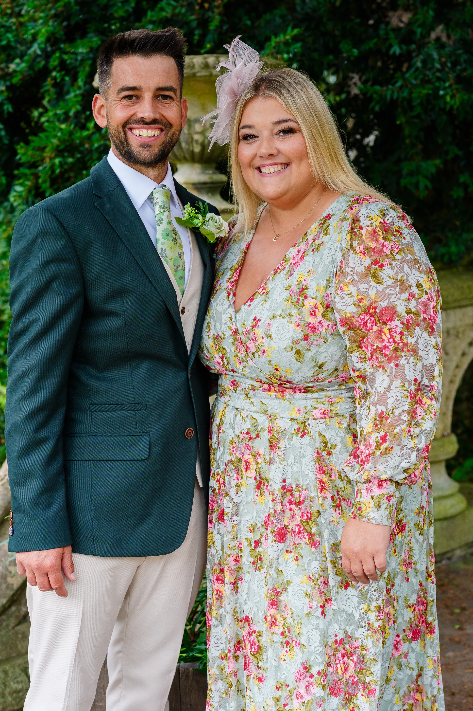A smiling couple posing outdoors with a green suit and a floral print dress against a leafy backdrop.
