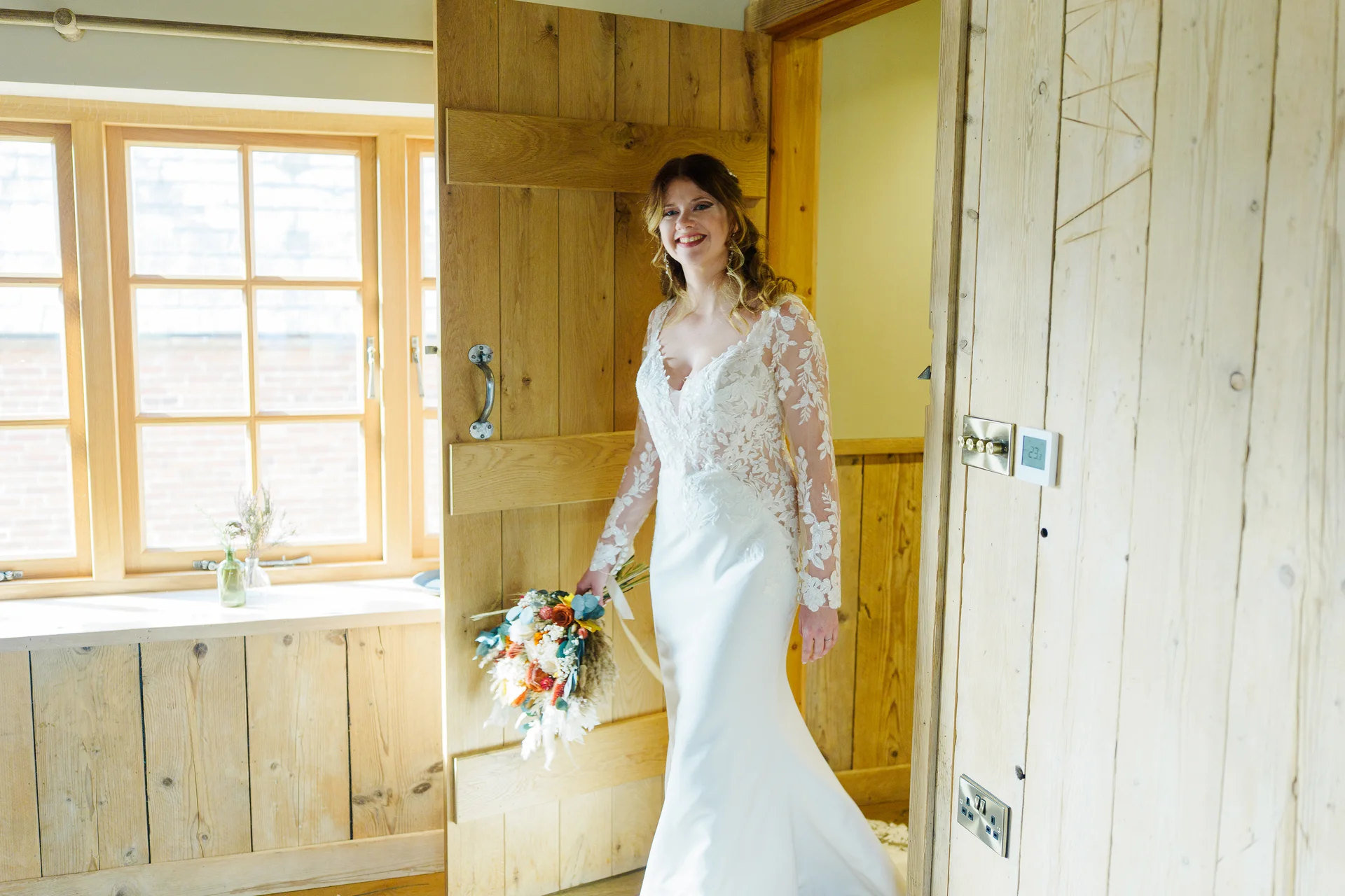 A smiling bride holding a colourful hand-tied bouquet poses in a rustic wooden room with large windows.