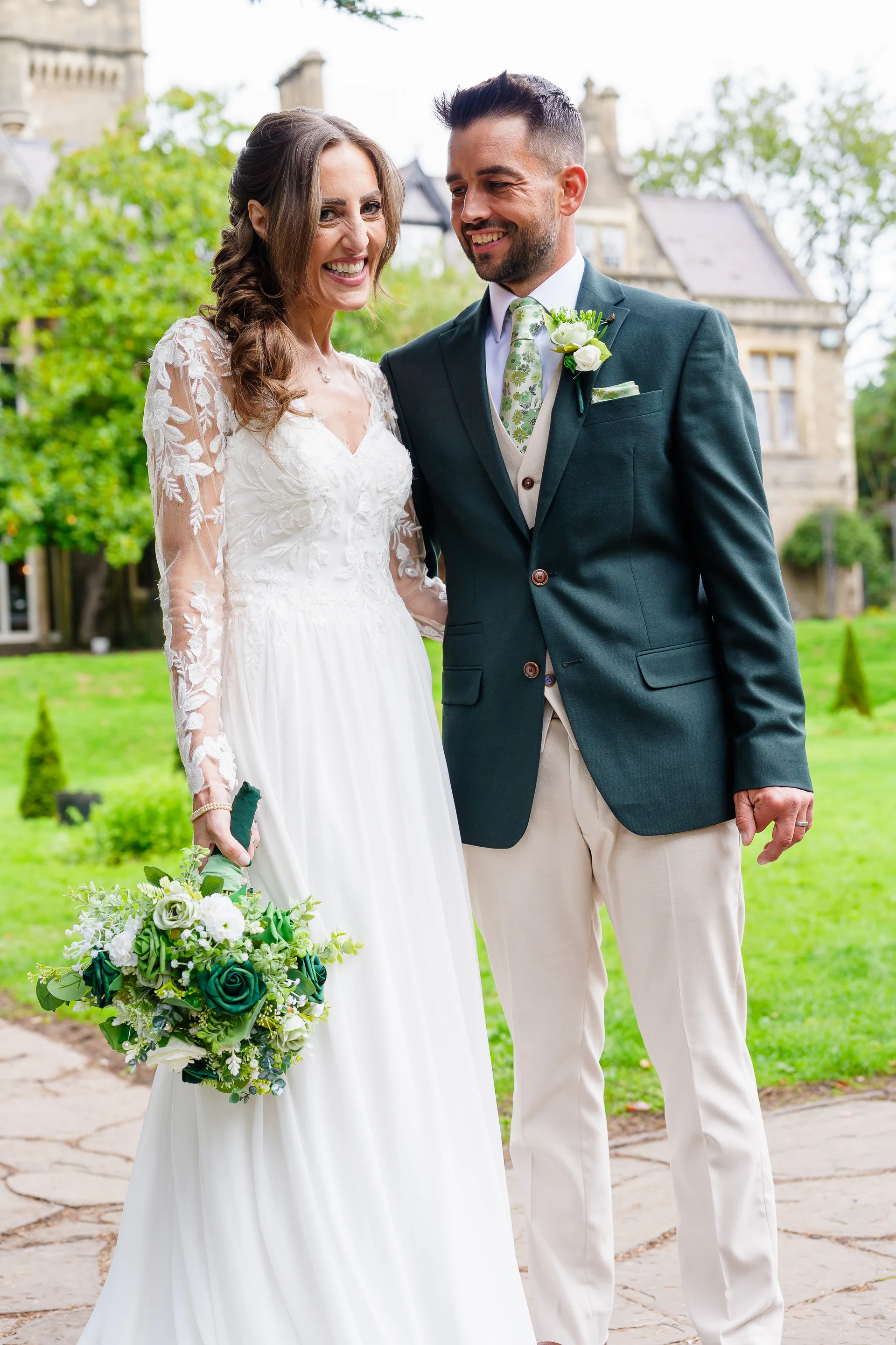 A smiling bride and groom pose in a lush manicured garden outside a grand stone manor house, holding a white and green bouquet.