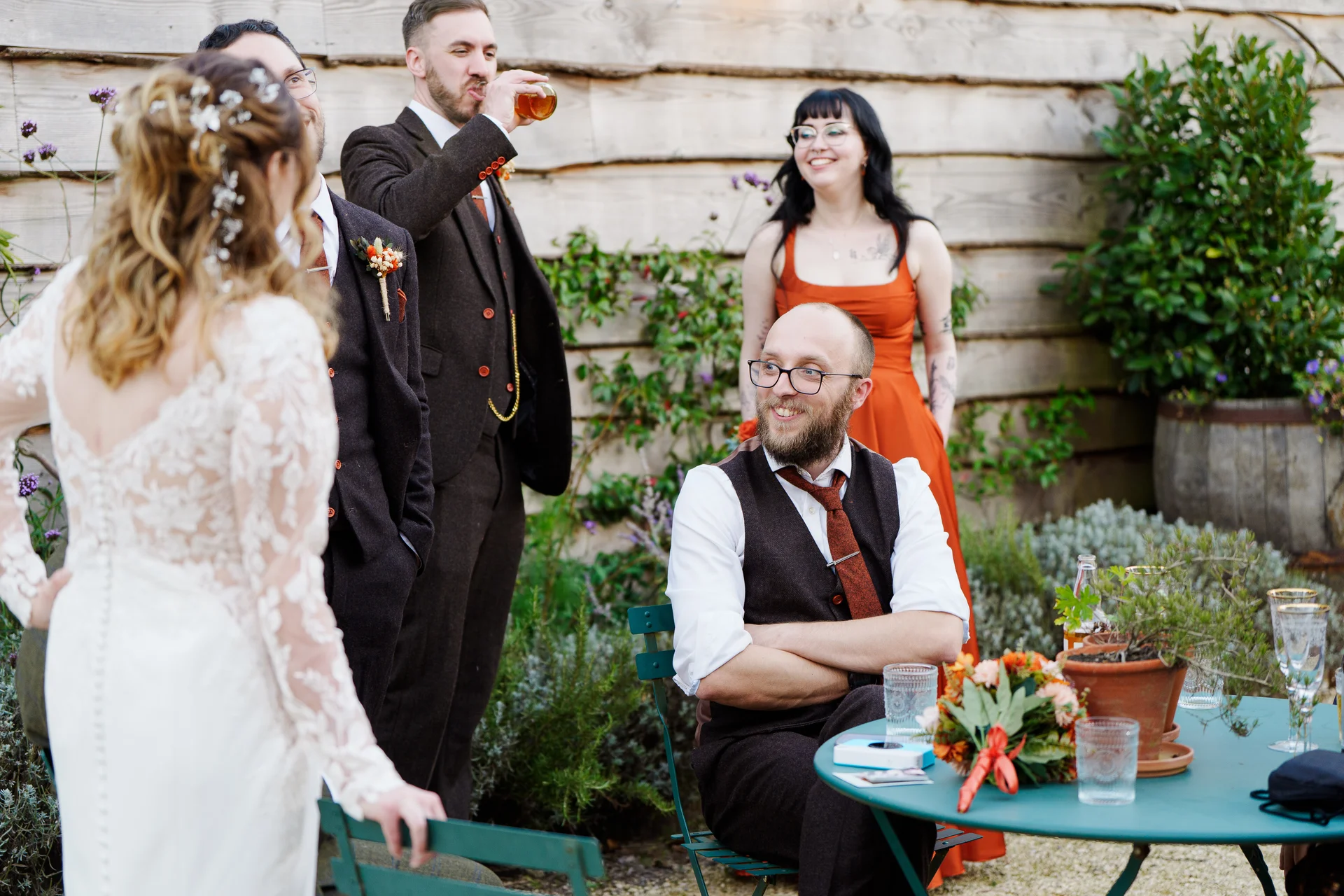 A bride in a lace gown chats with smiling wedding guests and groomsmen gathered outdoors near a rustic wooden wall.