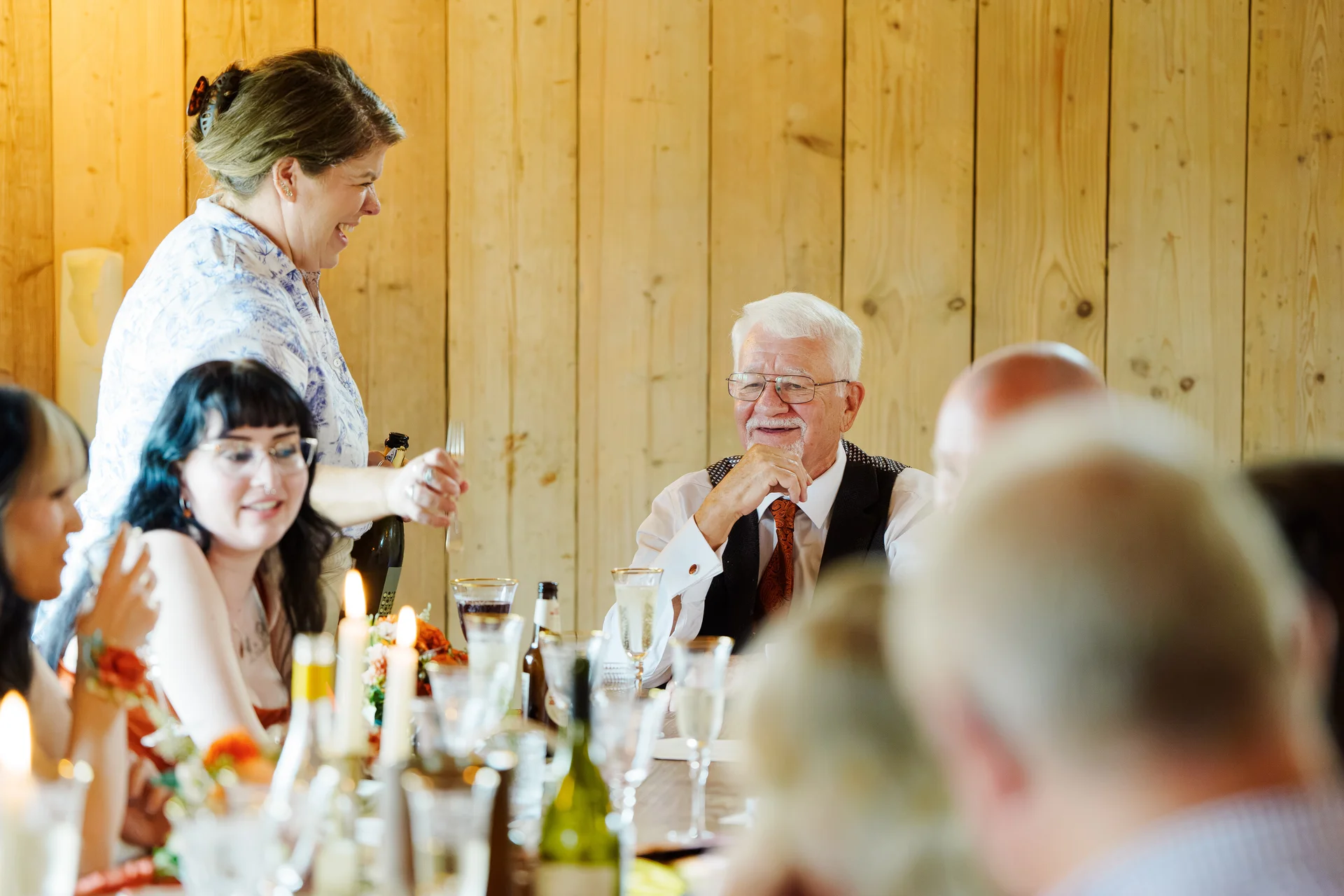 Guests enjoy lighthearted moments and laughter during the wedding breakfast seated at tables inside a rustic wooden barn venue.