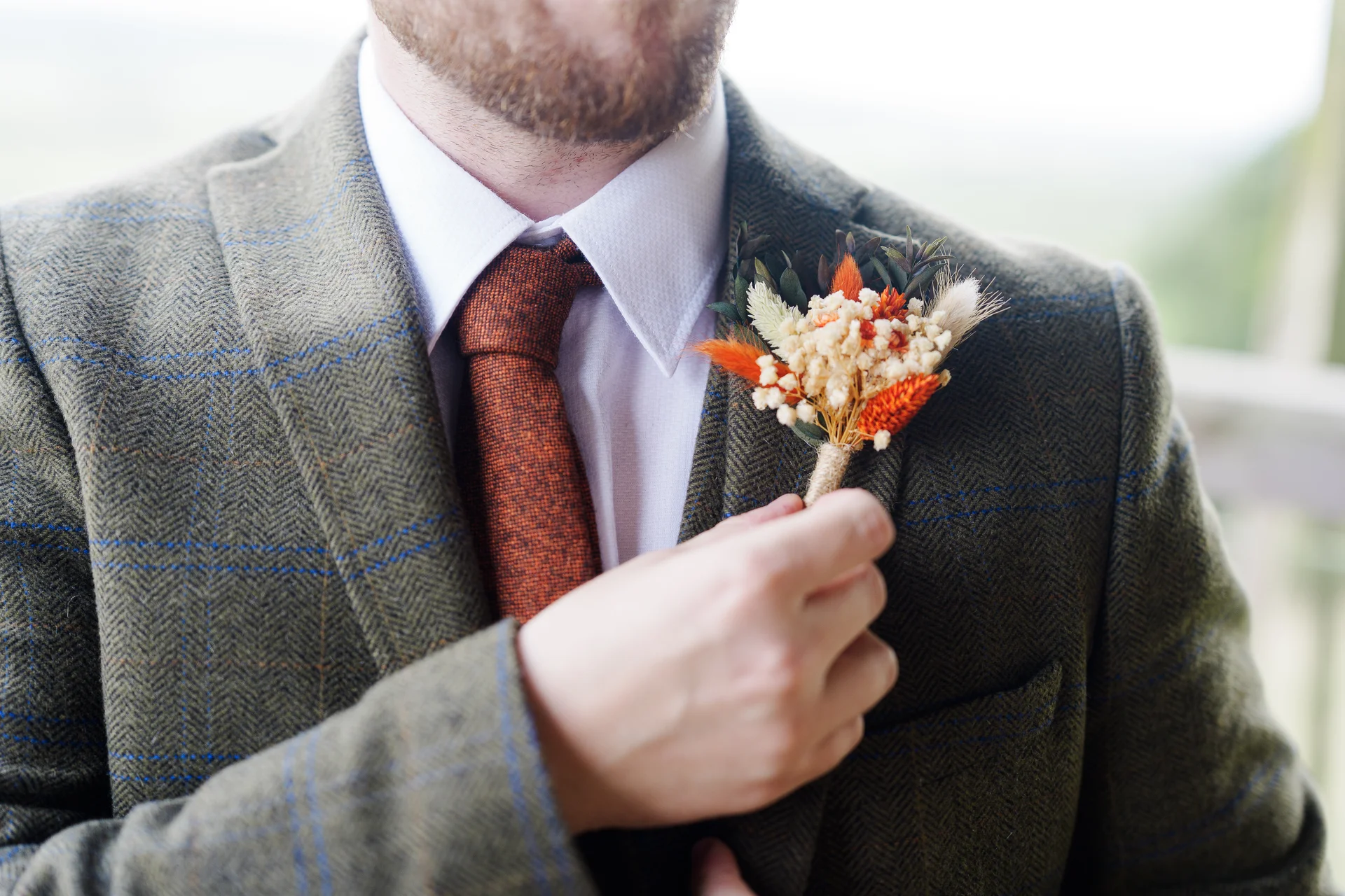 Close-up of a groom wearing a textured tweed suit and a burnt orange tie, adjusting a dried floral boutonniere pinned to his lapel.