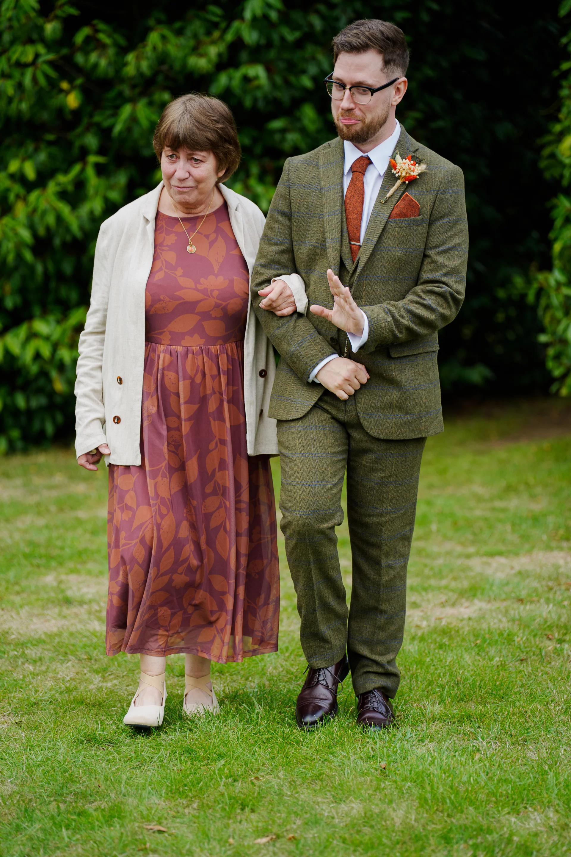 A groom in a green tweed suit walks arm in arm with his mother across a grassy lawn, both smiling softly during an outdoor wedding moment.