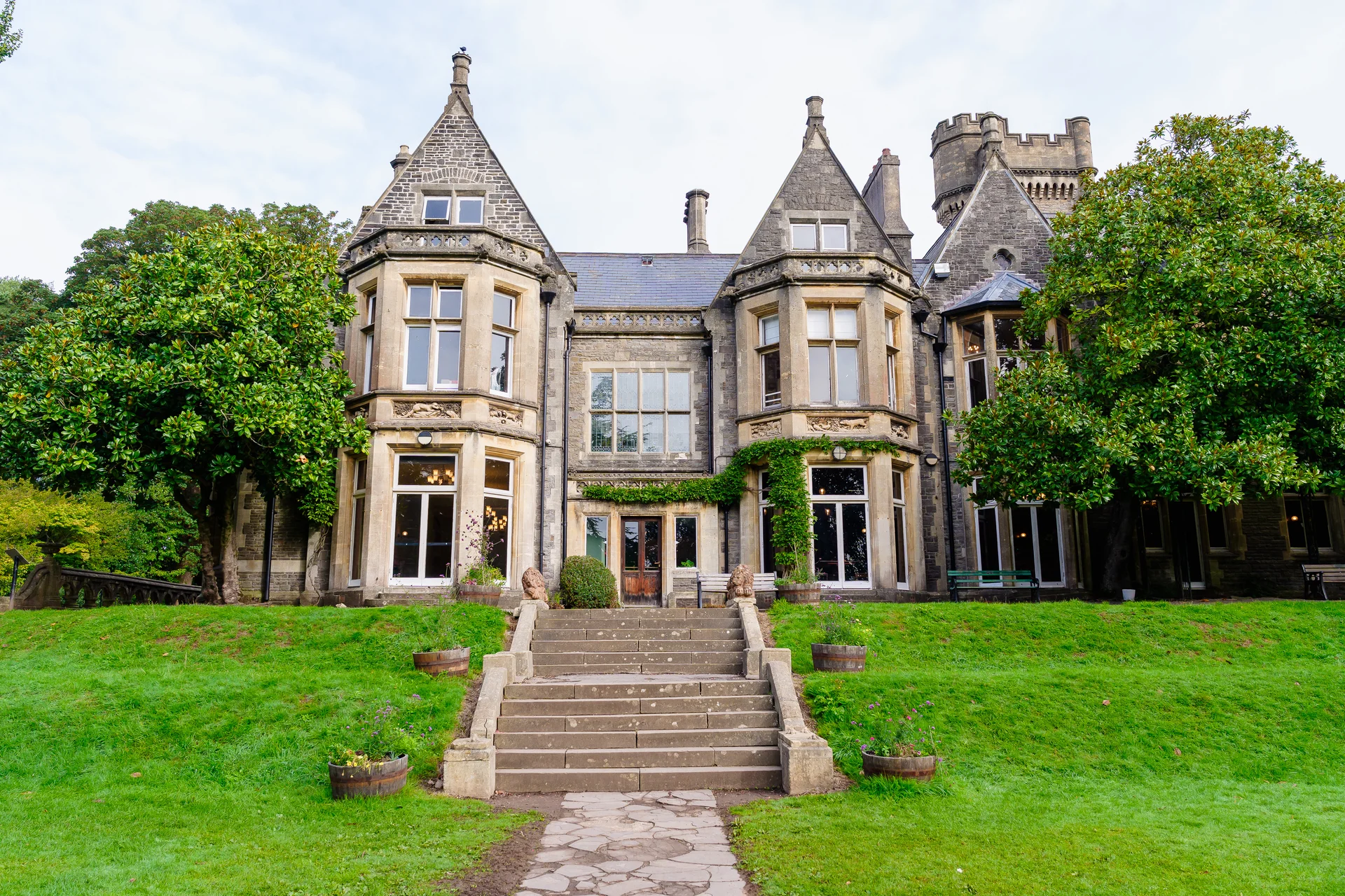 An exterior view of a historic stone castle wedding venue featuring grand bay windows, central stone steps, and lush green lawns.