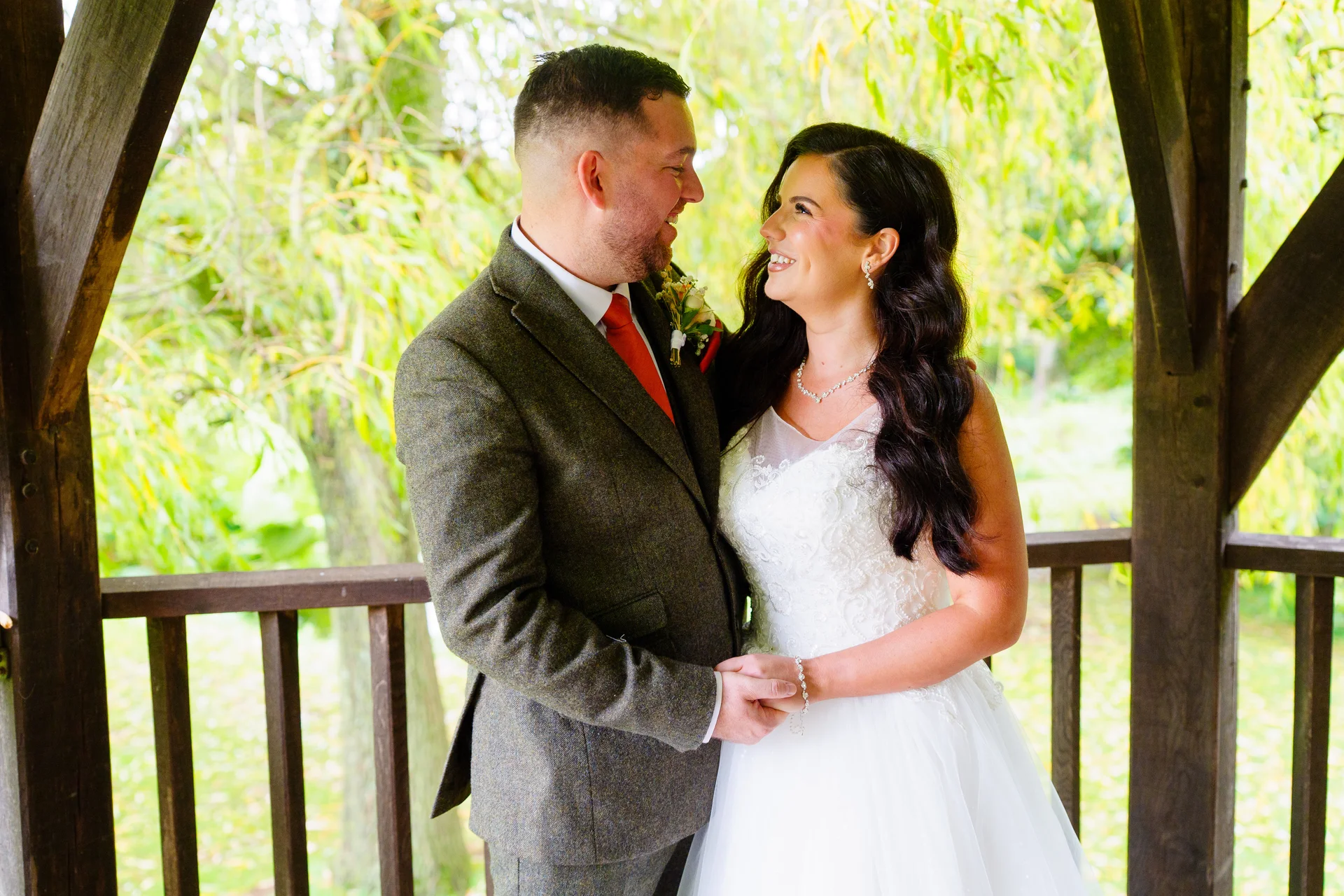 A smiling bride and groom gaze lovingly at each other while holding hands inside a wooden gazebo surrounded by lush greenery.