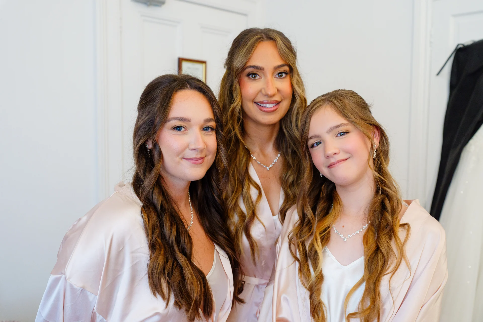 Three smiling women wearing matching pink satin robes and delicate necklaces pose together indoors during bridal morning preparations.