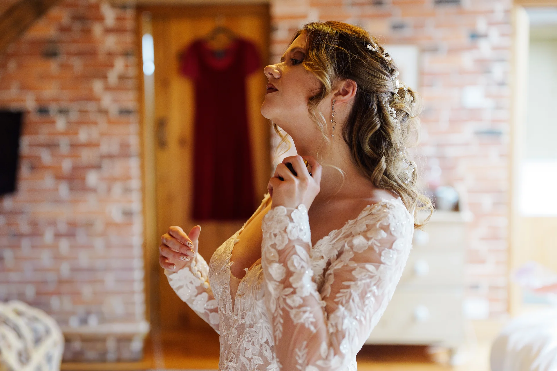 A bride in a lace gown with long sleeves applies perfume to her neck while getting ready against an exposed brick wall.
