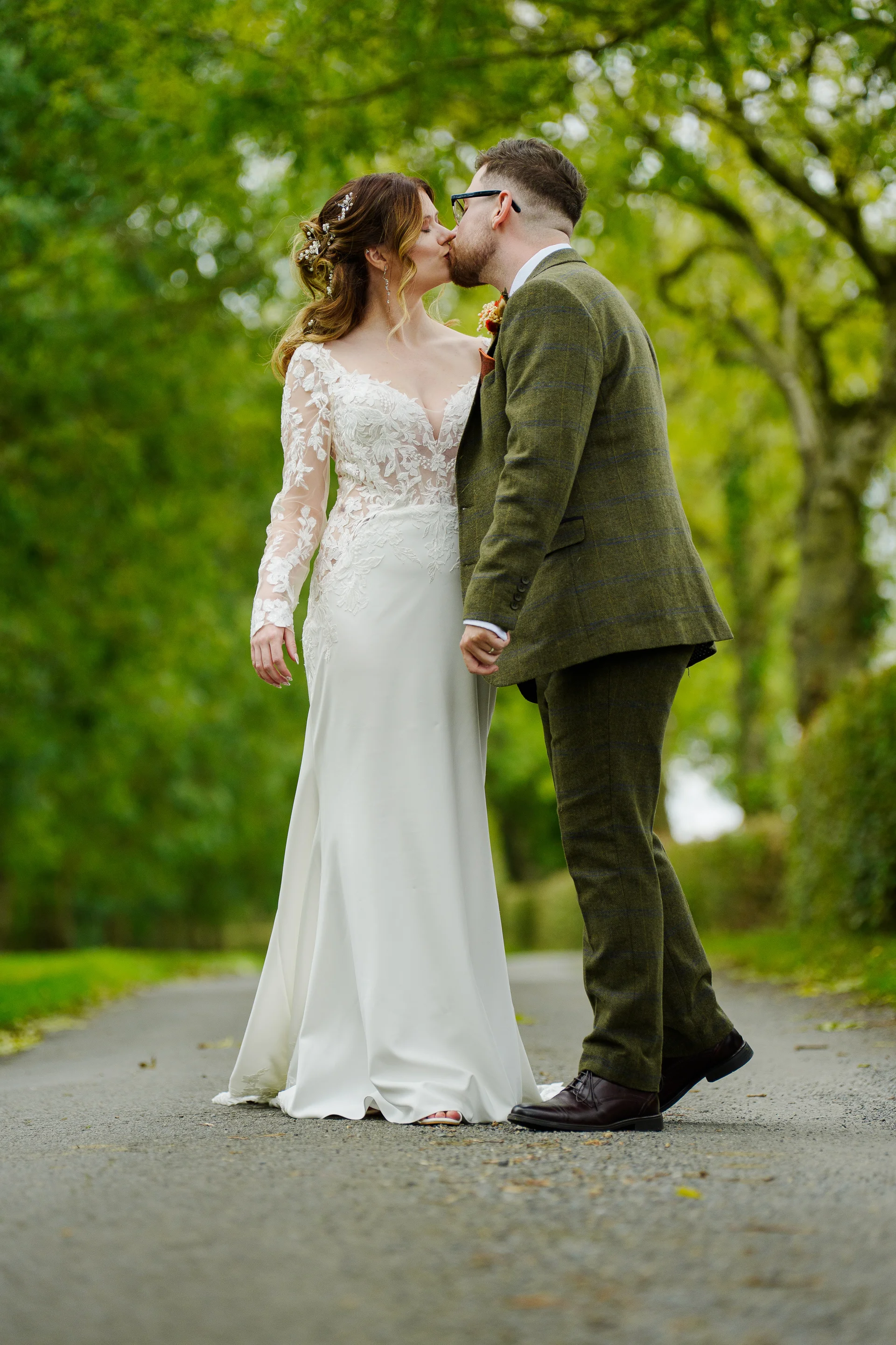 A newlywed couple shares a tender kiss on a paved pathway lined with lush green trees, the bride wearing a long-sleeved lace wedding gown and the groom in a green tweed suit.