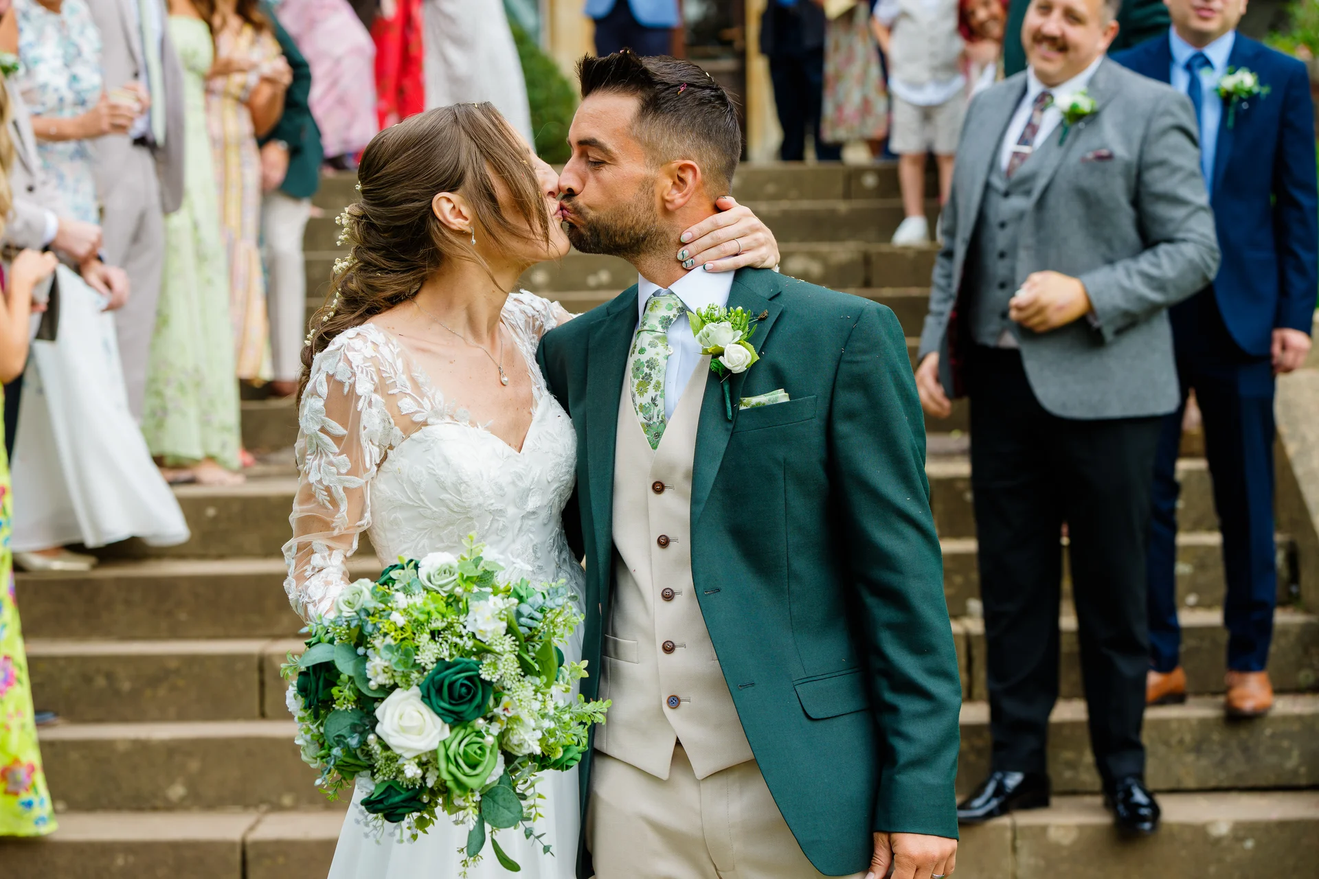 The bride and groom share a tender kiss on a grand outdoor stone staircase, holding a lush green-and-white bouquet while guests watch in the background.