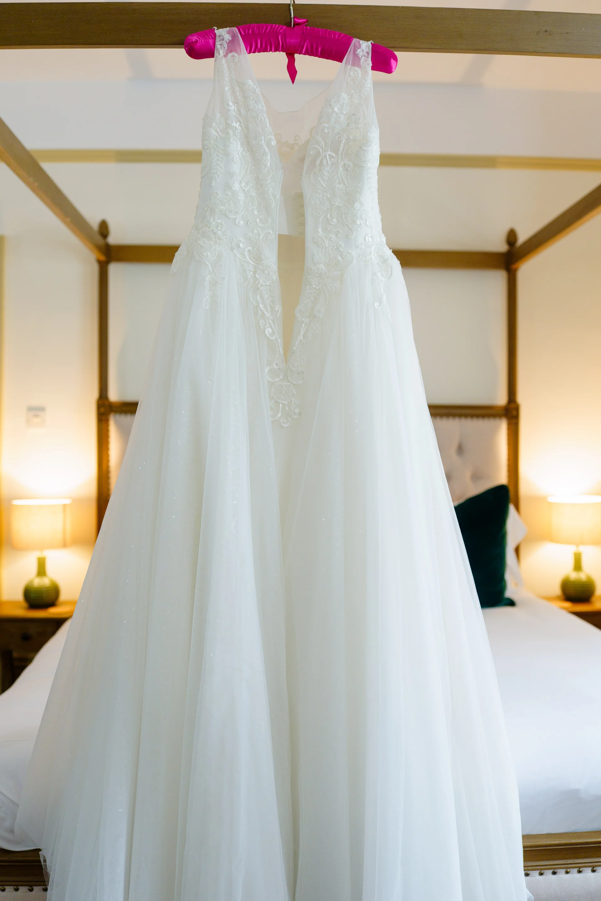 A detailed white wedding dress hangs on a bright pink padded hanger against a four-poster bed frame in a hotel room.