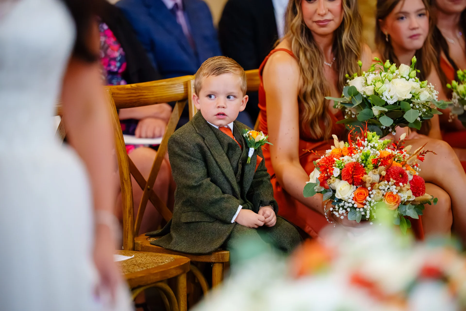 A young page boy in a smart green tweed suit sits attentively among seated wedding guests holding colourful bouquets.