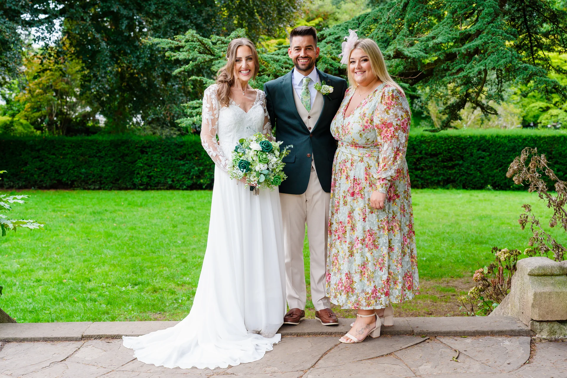 A smiling bride holding a bouquet of white and green roses stands next to a groom in a dark jacket and light trousers, alongside a female guest in a floral dress outdoors in a lush garden setting.