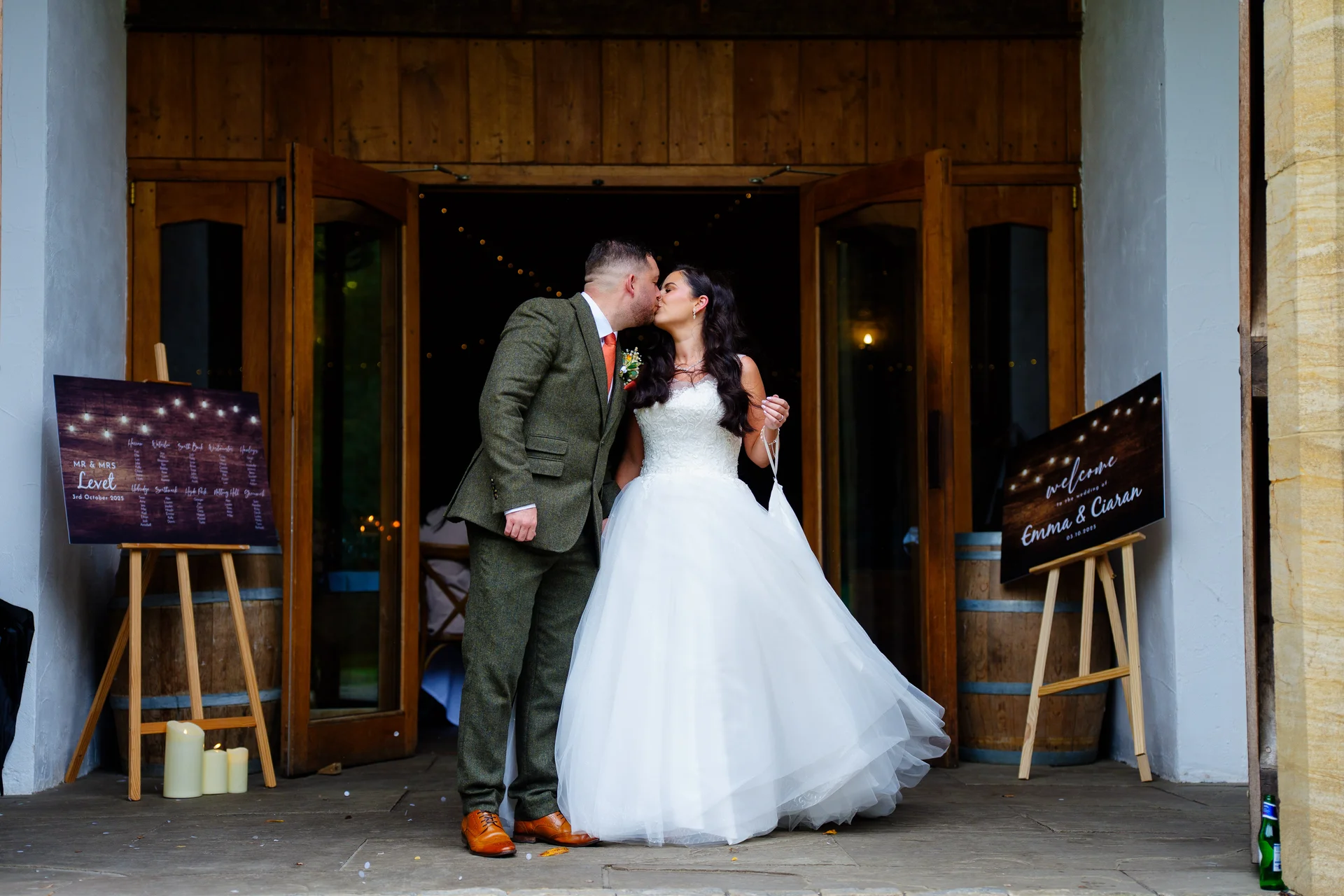 A newlywed couple shares a romantic kiss framed by the wooden double doors of a rustic barn venue, flanked by welcome signs on easels.