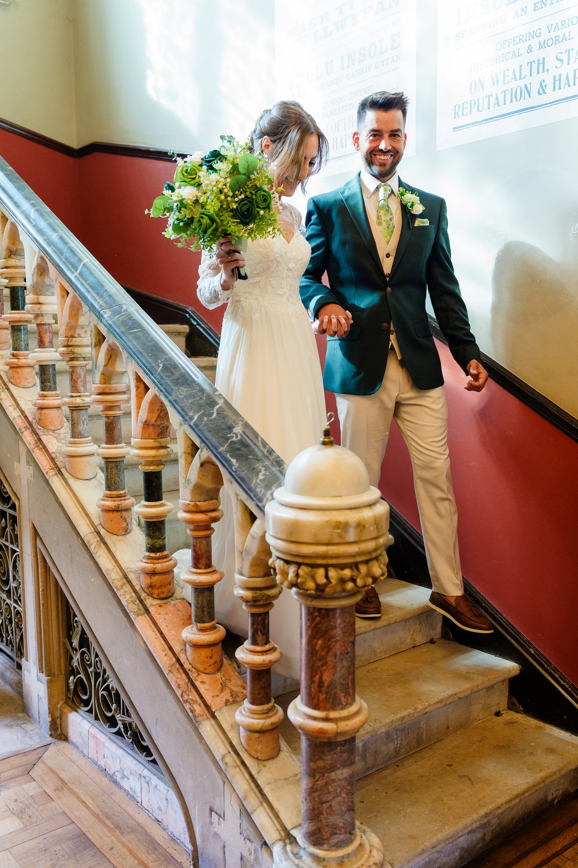 A smiling bride and groom holding hands walk down a grand stone staircase inside an elegant venue, with the bride holding a lush greenery bouquet.