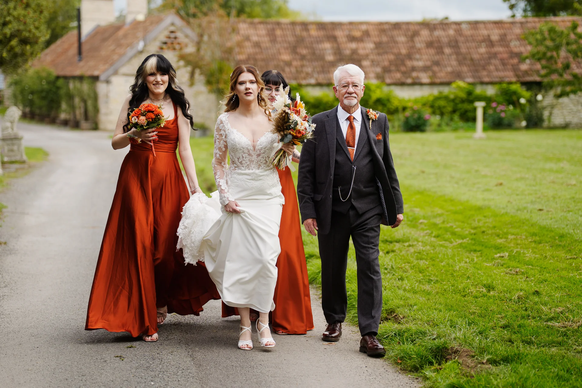A smiling bride wearing a lace-detailed long sleeve gown walks along a pathway alongside her father in a charcoal suit and bridesmaids in vibrant rust-orange satin dresses.