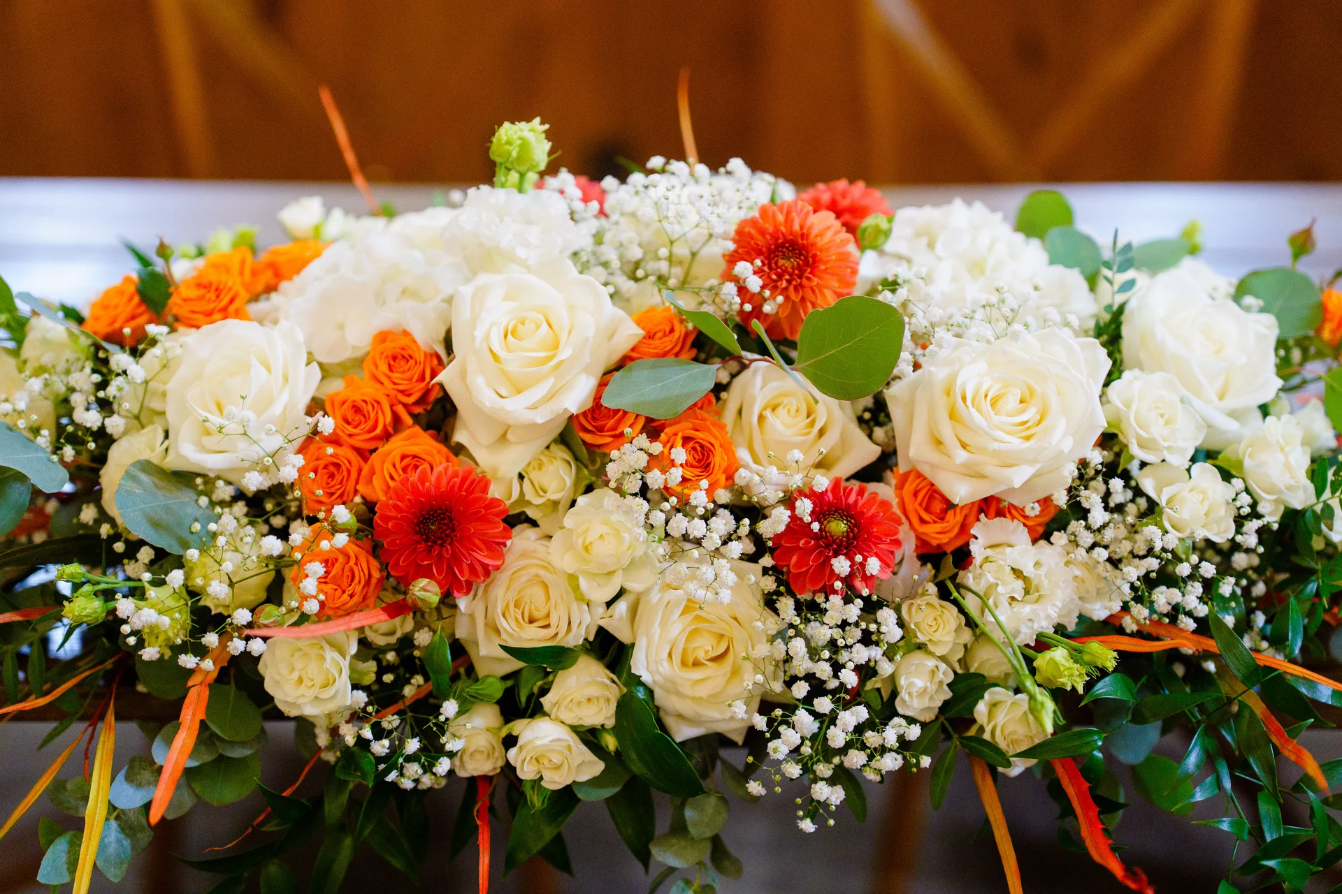 A close-up view of a vibrant top table floral arrangement featuring white roses, bright orange spray roses, chrysanthemums, and delicate baby's breath with eucalyptus foliage.