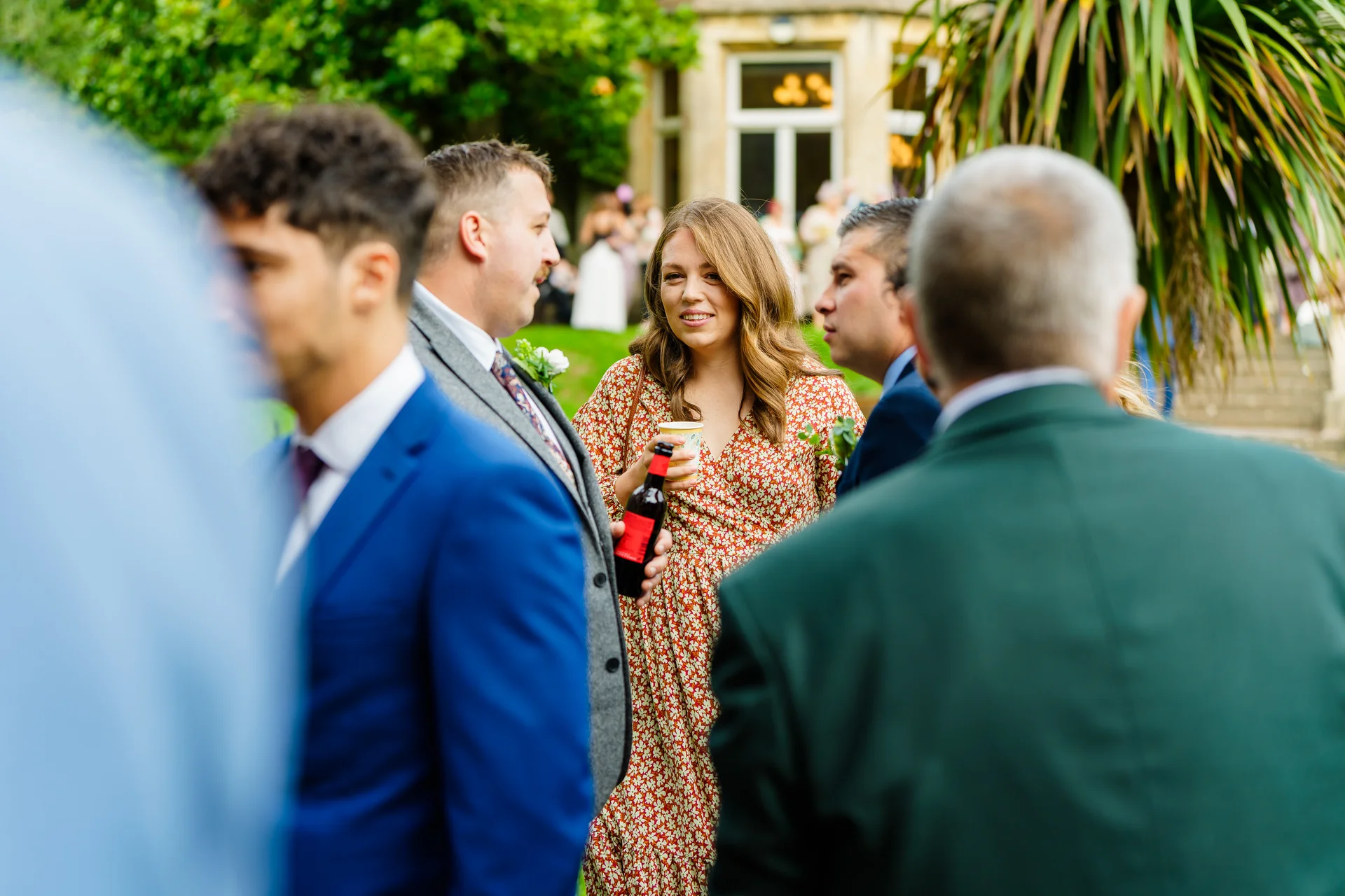 Wedding guests mingle and chat outdoors on a manicured lawn during a drinks reception in front of a grand manor house.