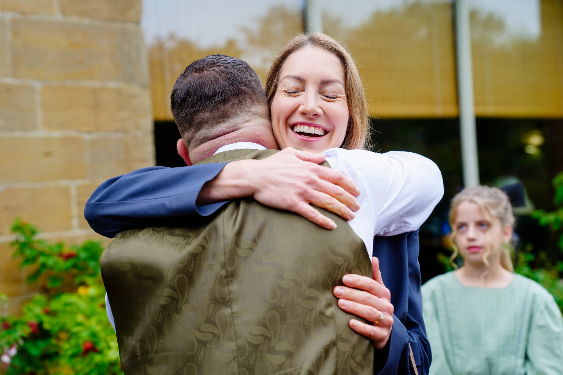 A joyful woman embraces a man wearing a patterned olive waistcoat while laughing happily during a wedding celebration.