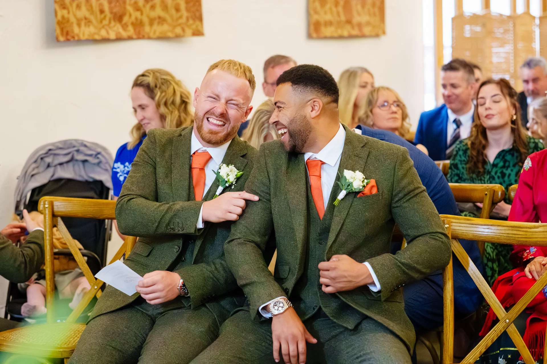 Two smiling grooms in matching green tweed suits and orange ties share a joyful laughing moment while seated among wedding guests.