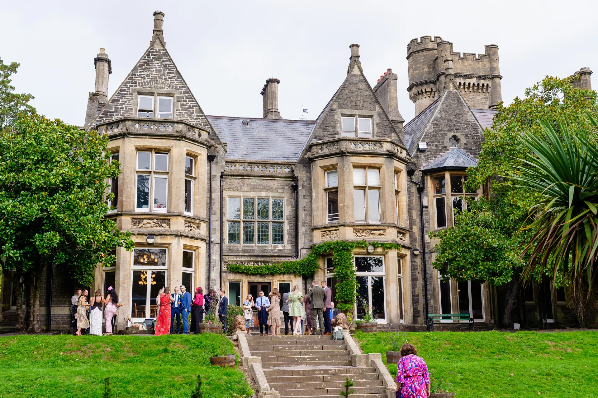 Wedding guests mingle on the lawn and stone steps in front of a grand historic castle venue under a soft overcast sky.