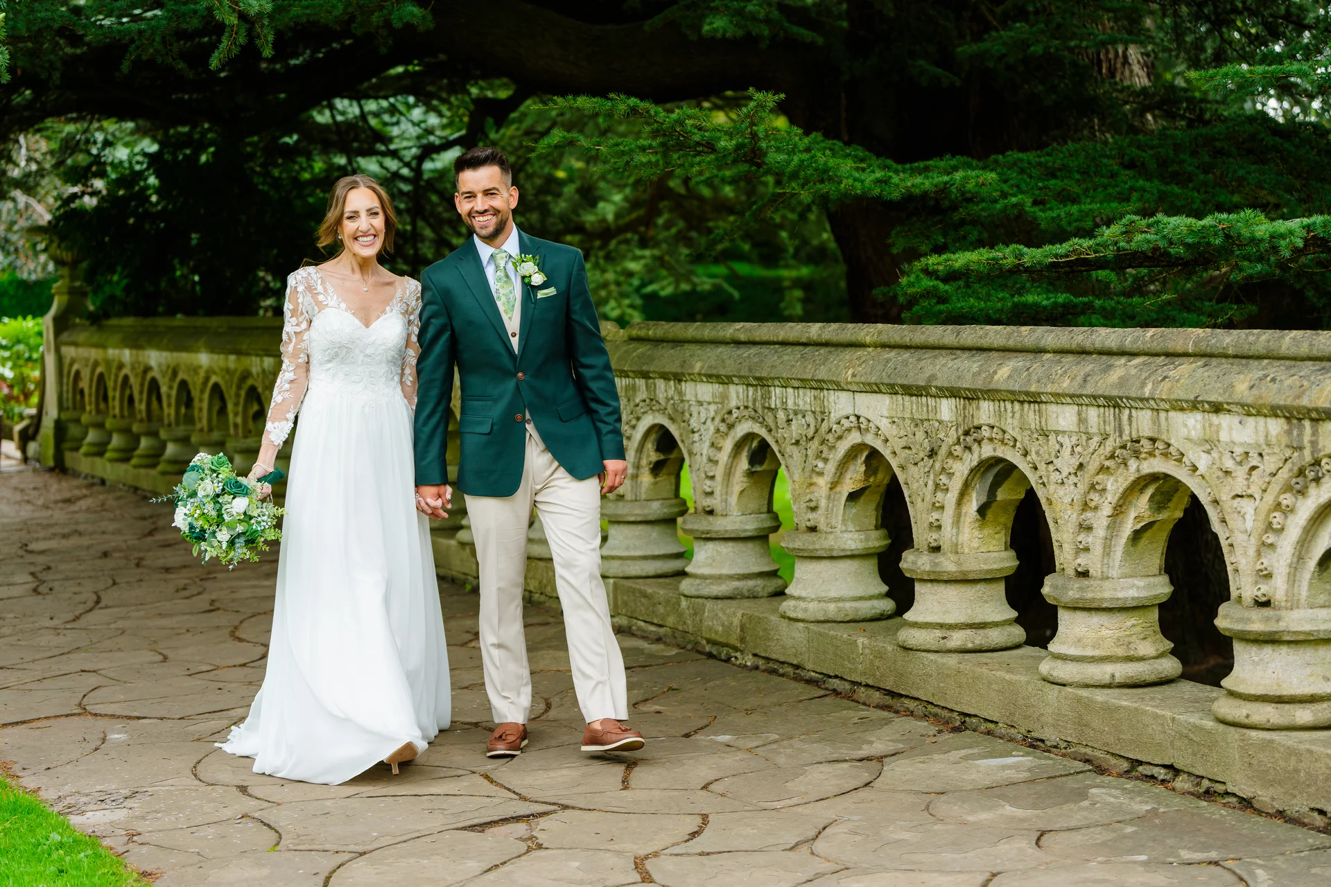 A smiling bride and groom walk hand in hand along an ornate stone bridge beside lush greenery on their wedding day.