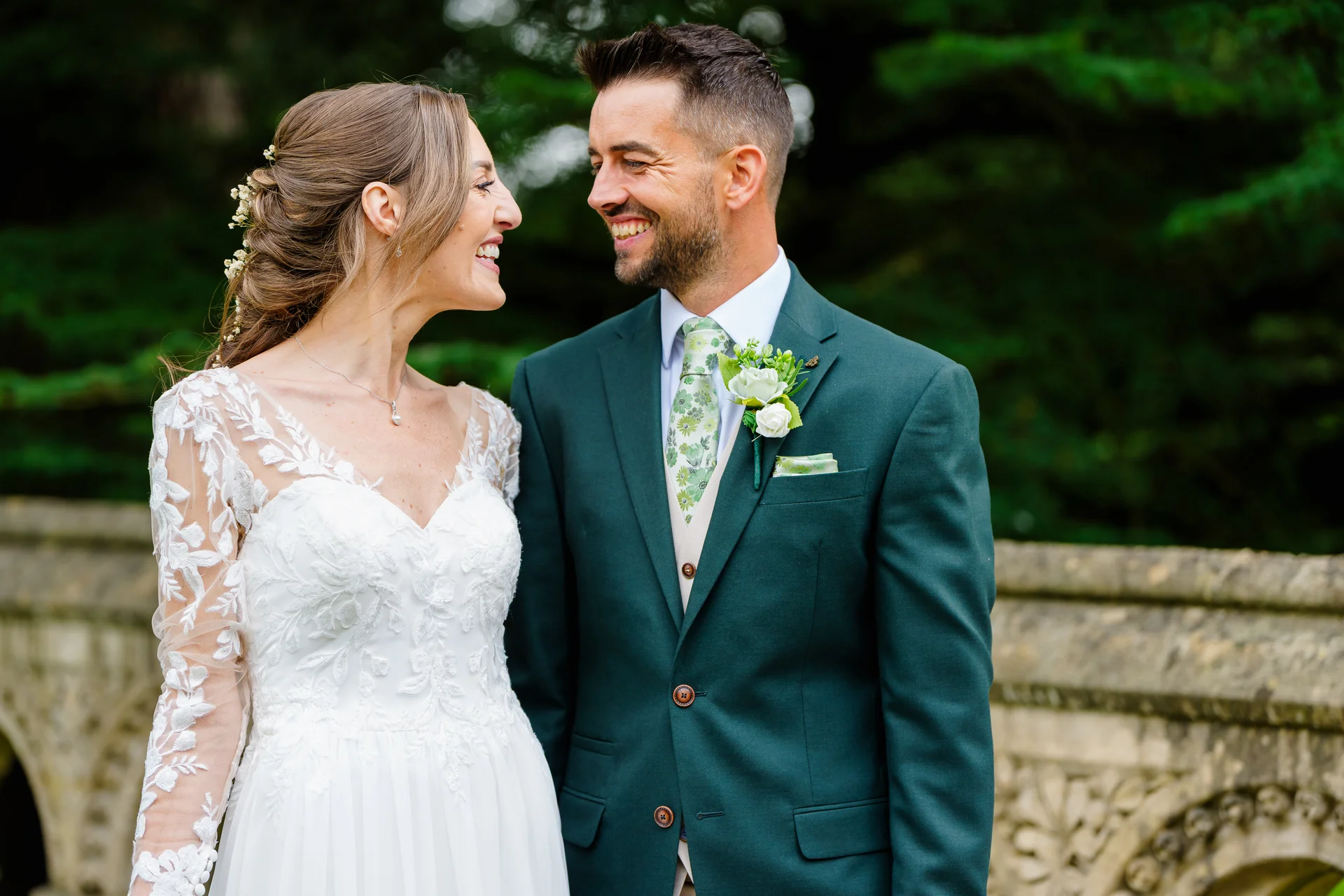 A smiling bride and groom gaze lovingly at each other outdoors, with the bride wearing a lace long-sleeve gown and the groom in a forest green suit.