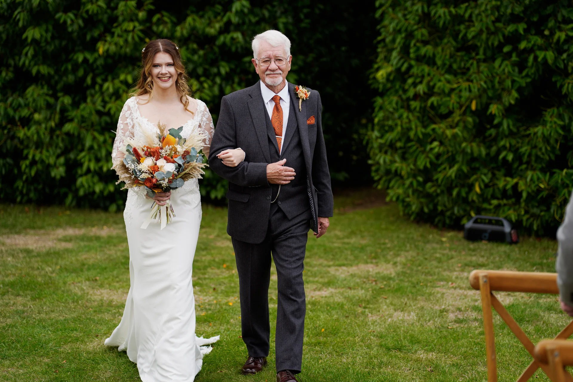 A smiling bride walks down the grass aisle arm-in-arm with her father, holding a colourful autumnal bouquet featuring dried grasses and orange florals.