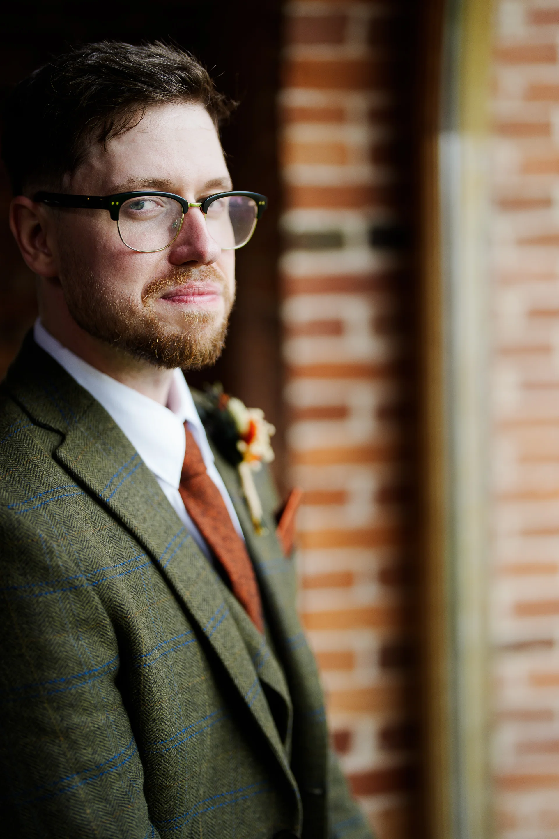 A handsome groom wearing a green tweed suit, orange tie, and glasses stands against an exposed brick wall, looking thoughtfully over his shoulder.