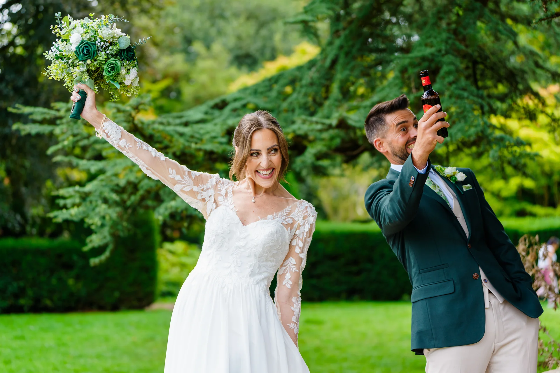 A joyful bride in a lace long-sleeve gown raises her bouquet while her groom in a green suit playfully raises a beer bottle outdoors against a lush green garden backdrop.