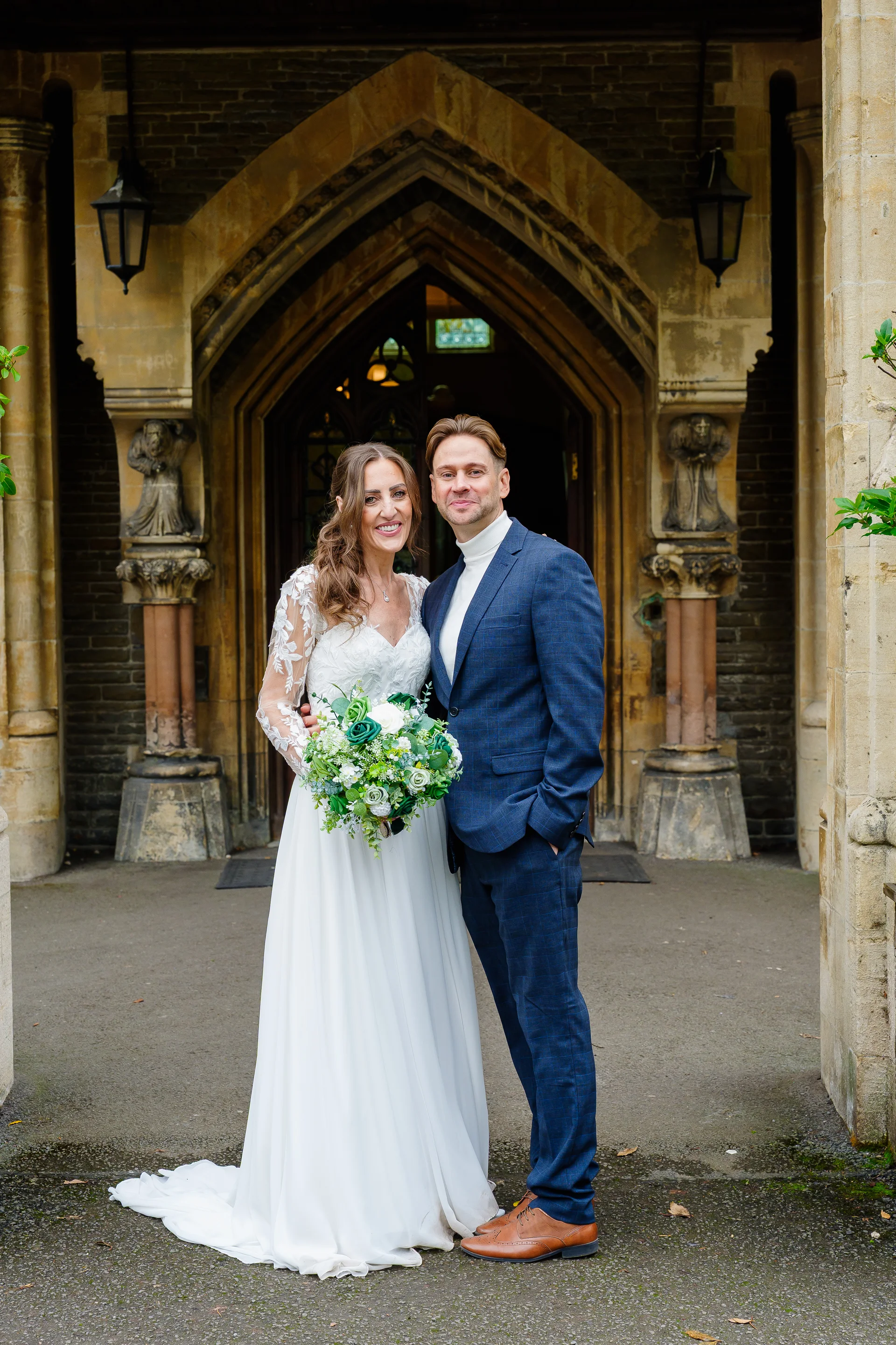 A smiling bride and groom pose together outside a historic stone church entrance, with the bride holding a lush white and green bouquet.