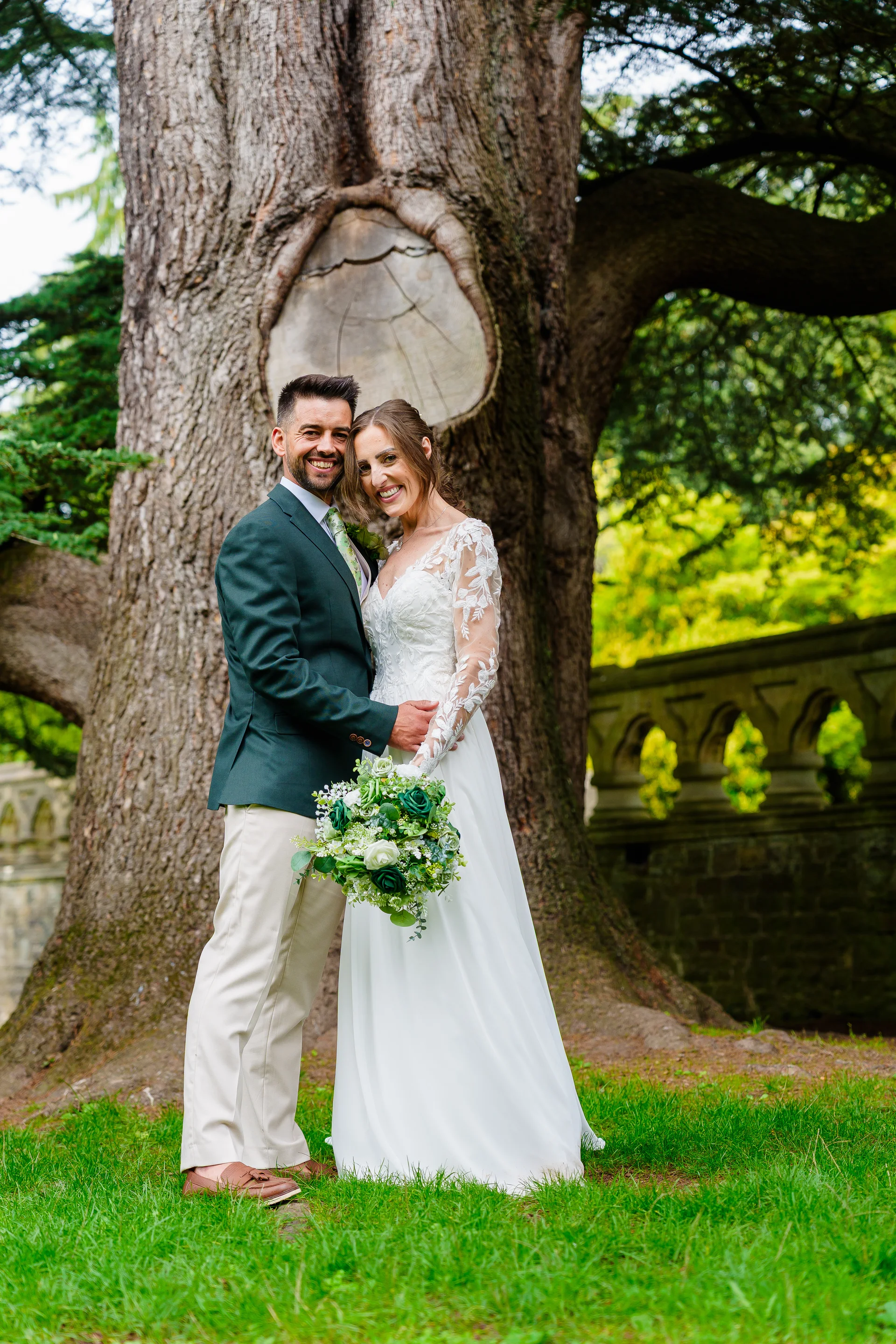 Newlywed couple smiling warmly together outdoors, standing before a massive ancient oak tree while the bride holds a lush green and white bouquet.