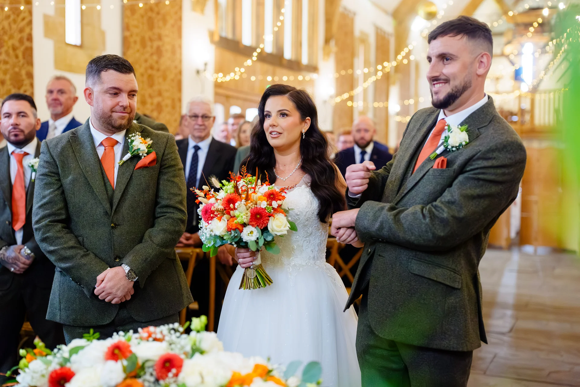 A smiling bride holding a vibrant orange and white bouquet stands between the groom and a groomsman at the altar inside a rustic oak-framed barn.