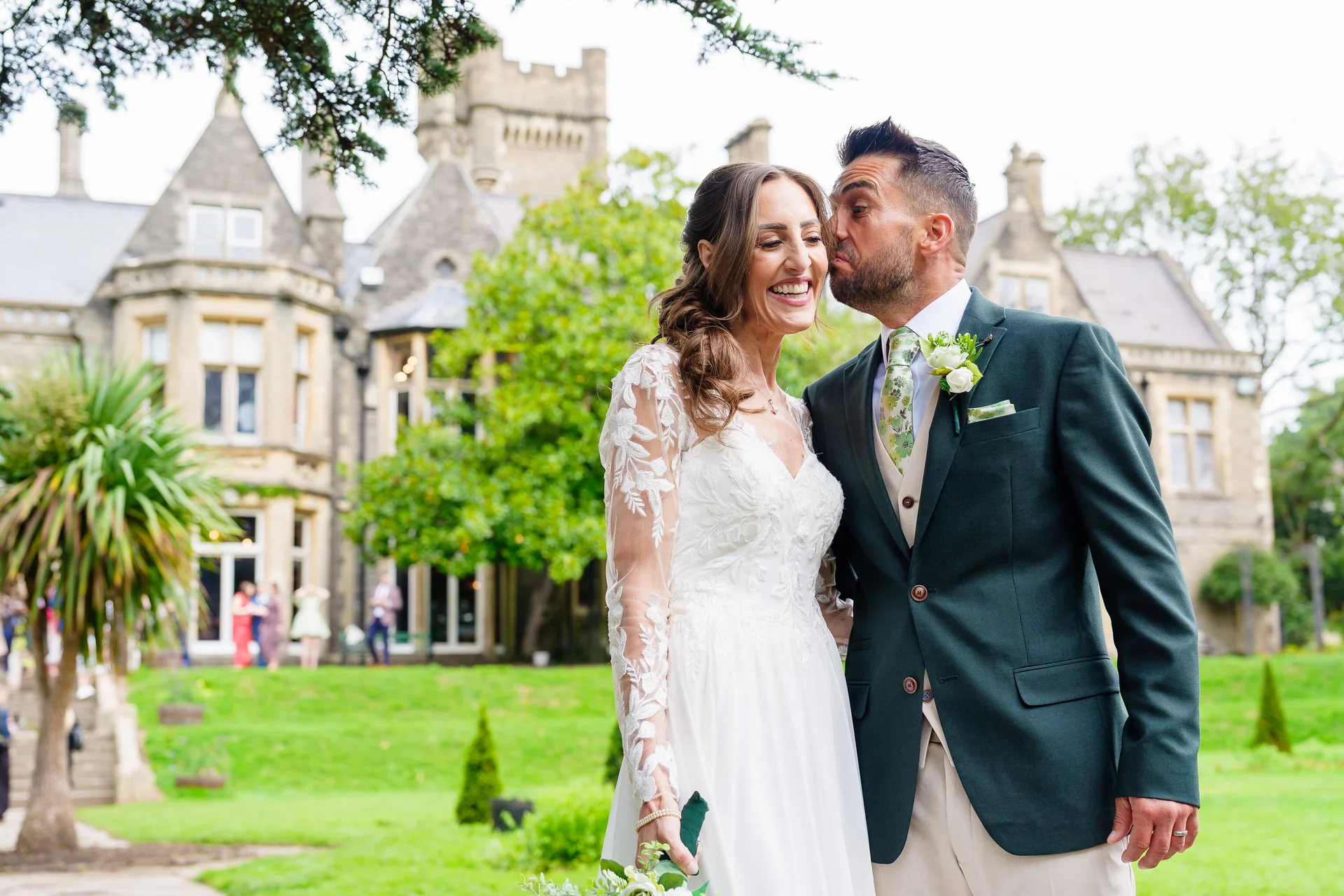 A joyful newlywed couple shares a playful moment outside a grand country house, with the groom leaning in to kiss the laughing bride's cheek.