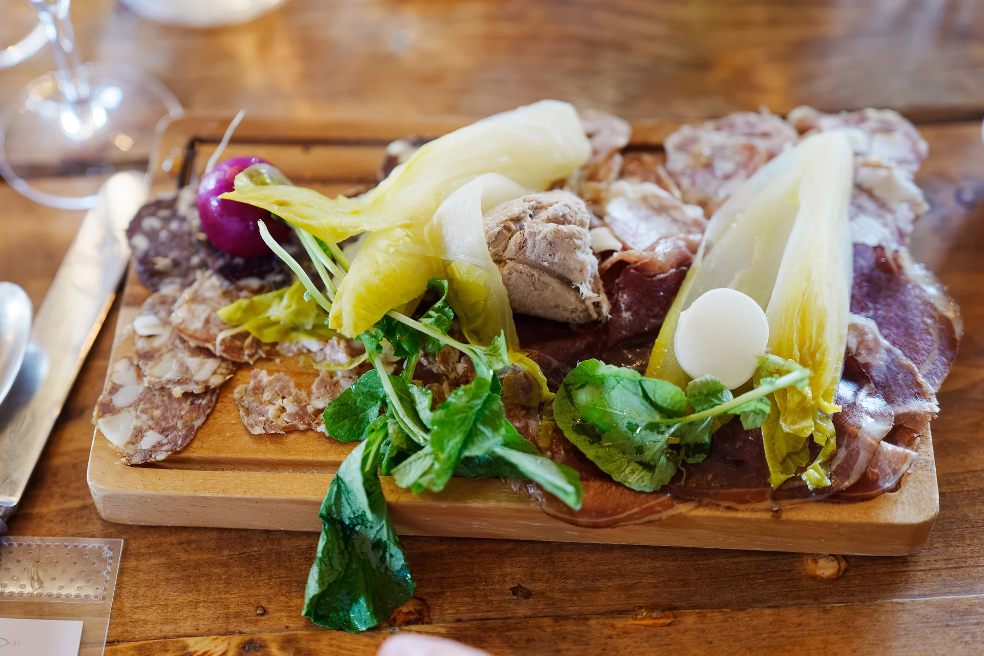 A rustic wooden sharing board laden with cured meats, pate, fresh salad leaves, and pickled vegetables sits on a wooden table.