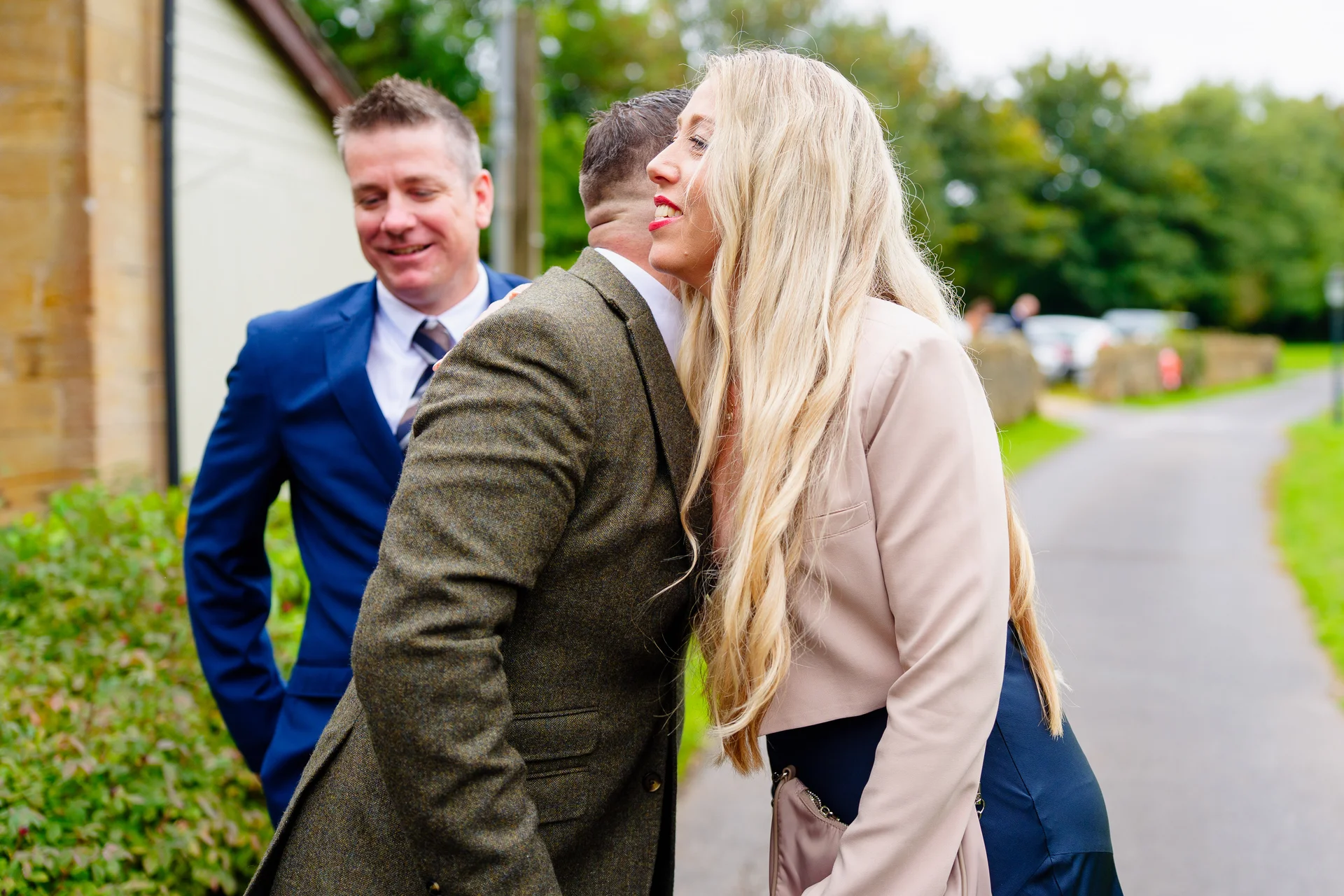 A smiling blonde female wedding guest greets another guest with a warm hug outdoors while a man in a blue suit looks on happily.