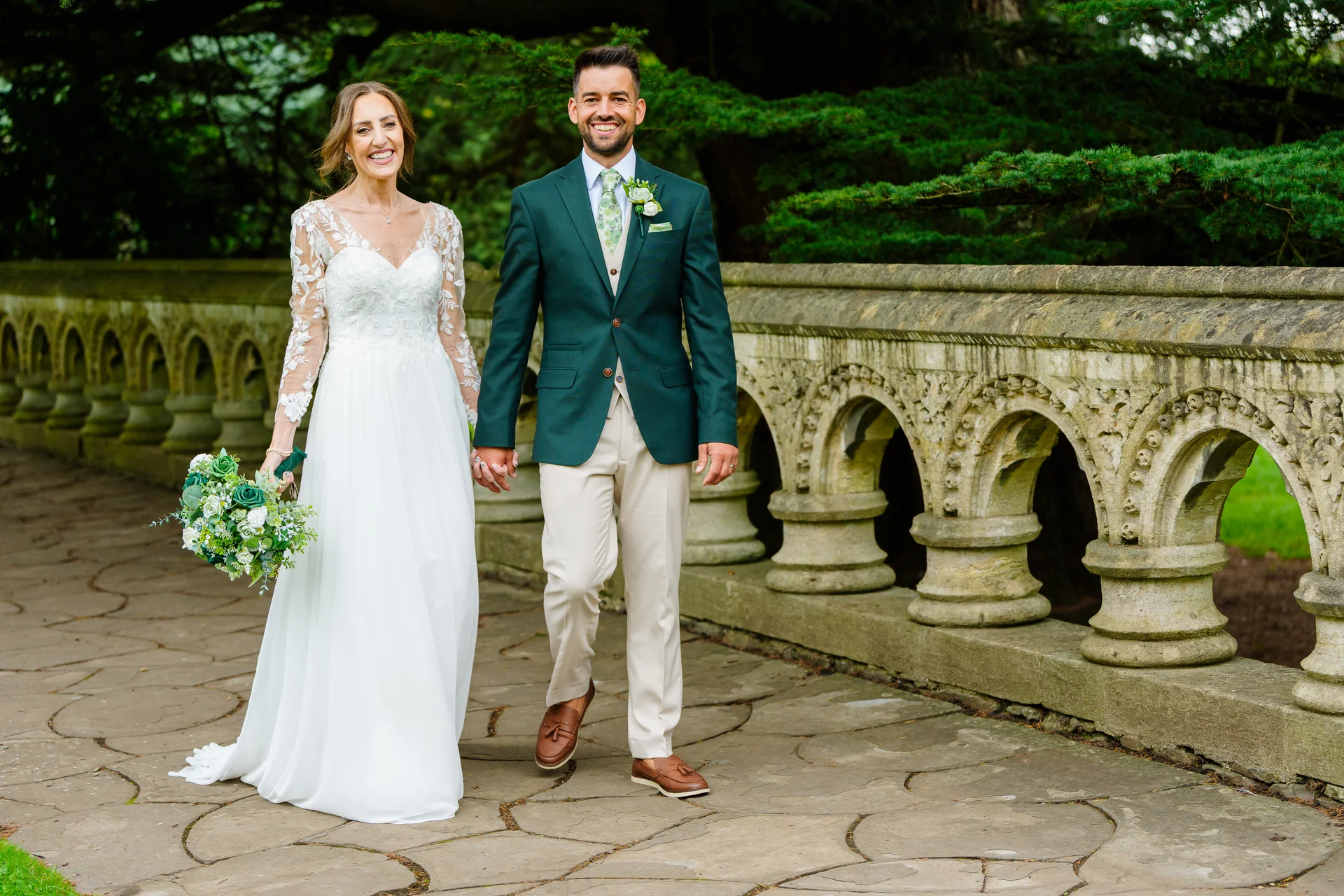 A smiling bride holding a green bouquet and groom in a dark green jacket walk hand in hand along a stone path by an ornate balustrade.