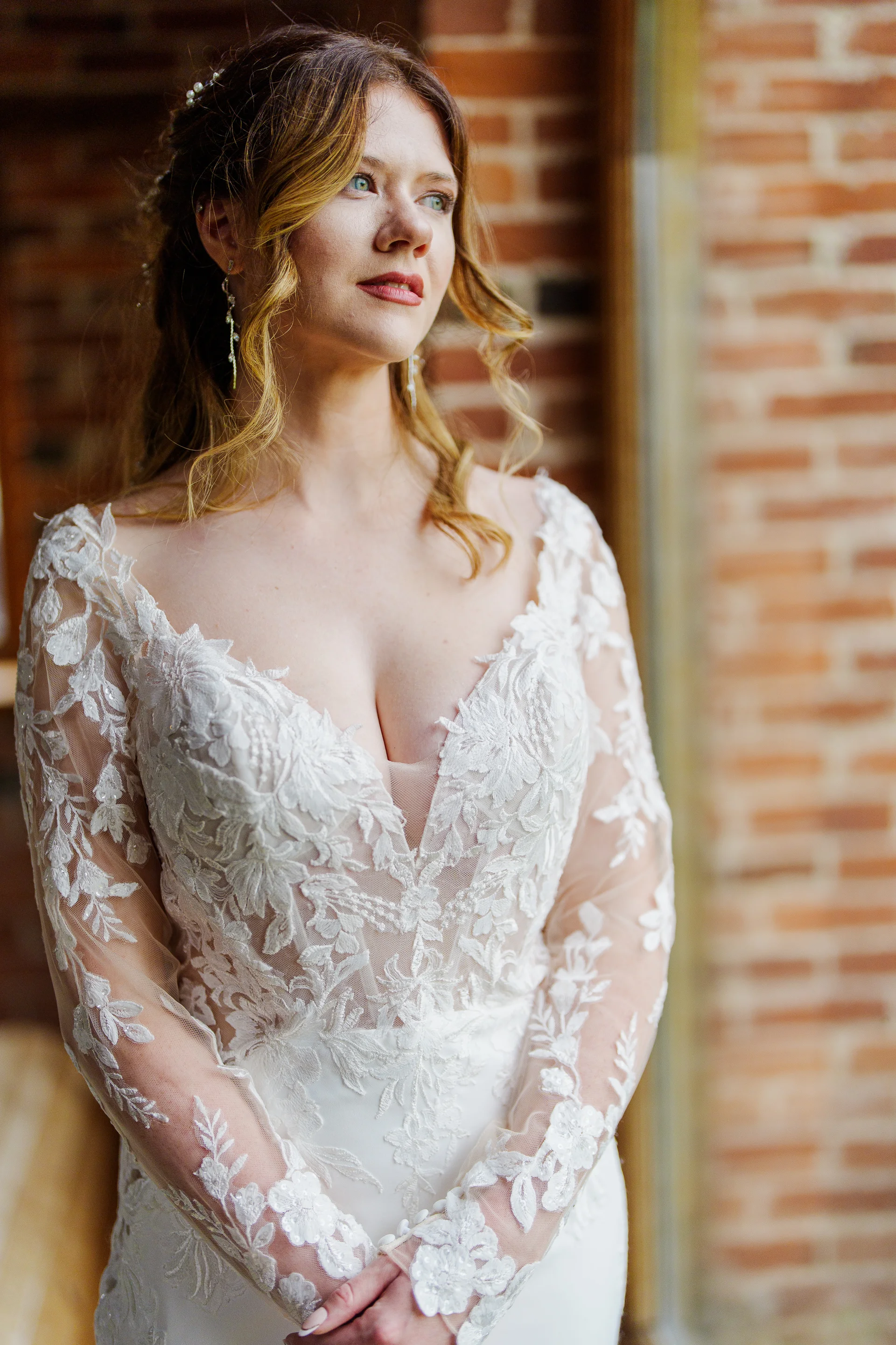 A stunning bride stands elegantly against an exposed brick wall, wearing a long-sleeved lace wedding dress with a plunging neckline.
