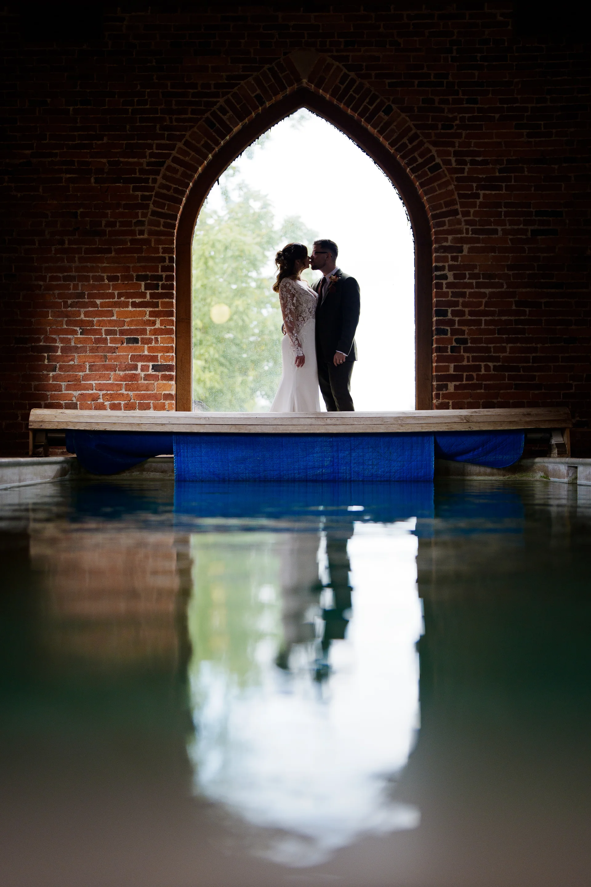 A bride and groom share a tender kiss framed inside a large stone archway with a stunning reflection of them shimmering in the water below.