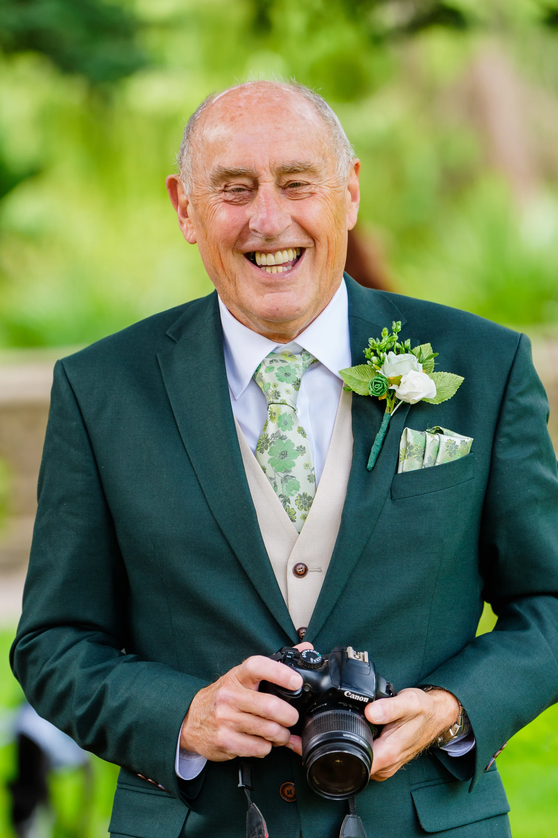 An older guest wearing a dark green suit, beige waistcoat, and floral tie laughs joyfully while holding a camera outdoors.