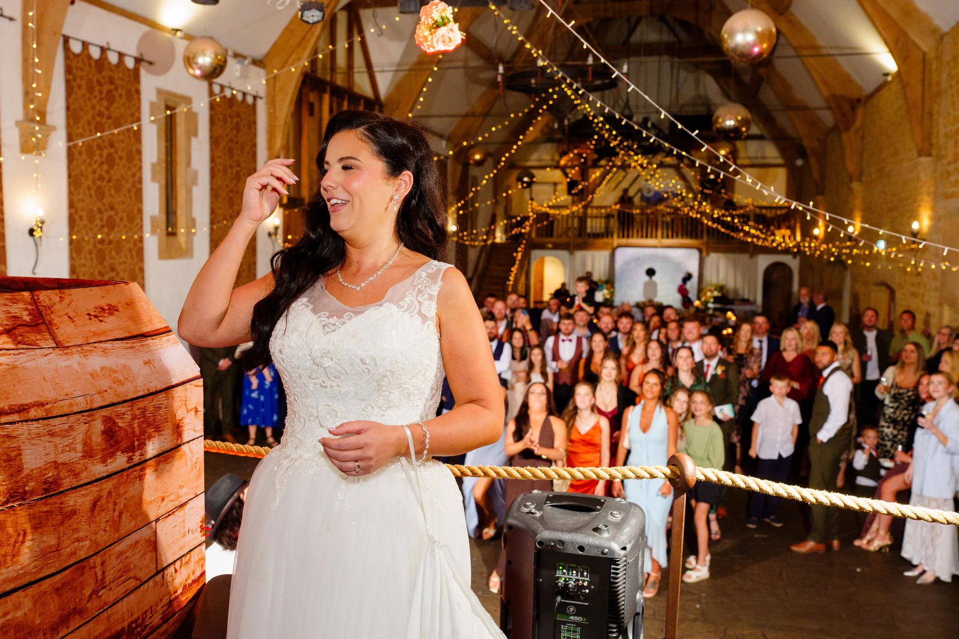 A smiling bride delivers a speech wearing a lace wedding dress in a rustic stone barn illuminated by fairy lights and disco balls.