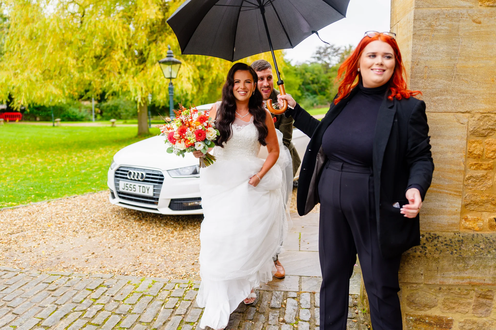 A joyful bride holding a colourful bouquet walks out into the rain under a black umbrella, accompanied by her groom and a coordinator on a cobbled pathway beside a white wedding car.