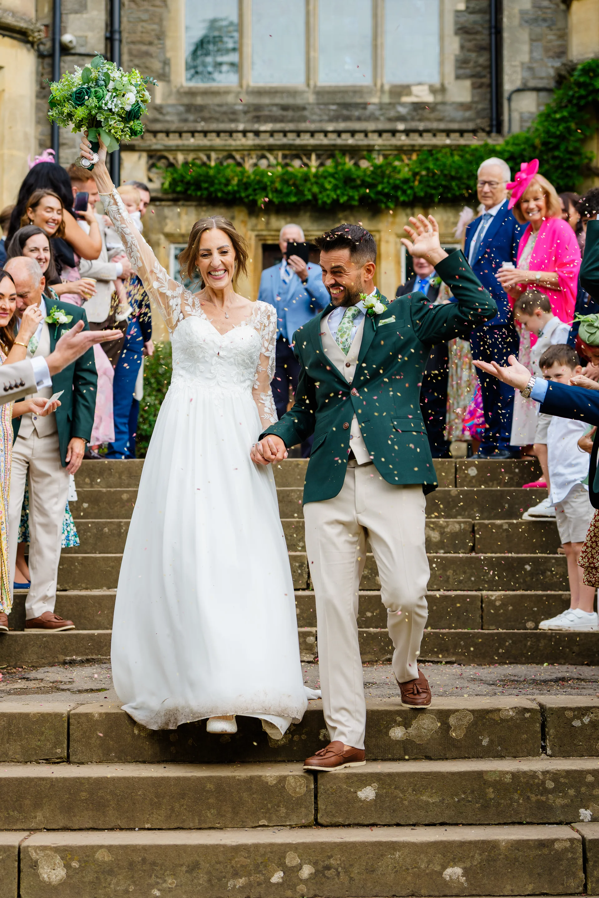 A bride in a long-sleeved lace dress and a groom in a green suit walk down stone steps while guests throw colourful confetti during their wedding celebration.