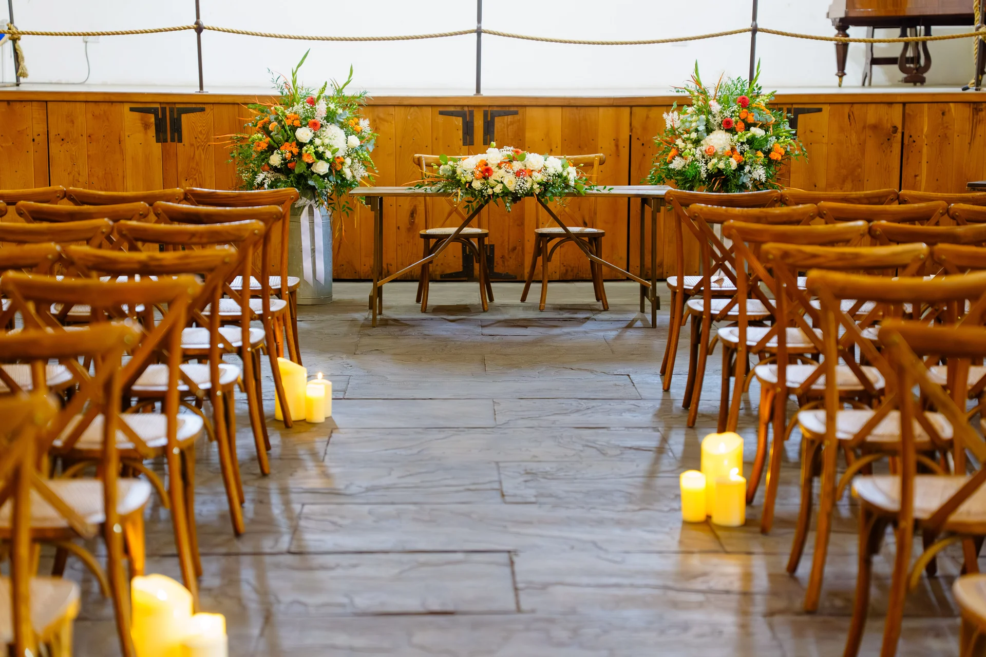 A wide view of a rustic wedding ceremony setup featuring a sweetheart table adorned with floral arrangements and flanked by rows of wooden chairs and glowing pillar candles along the aisle.