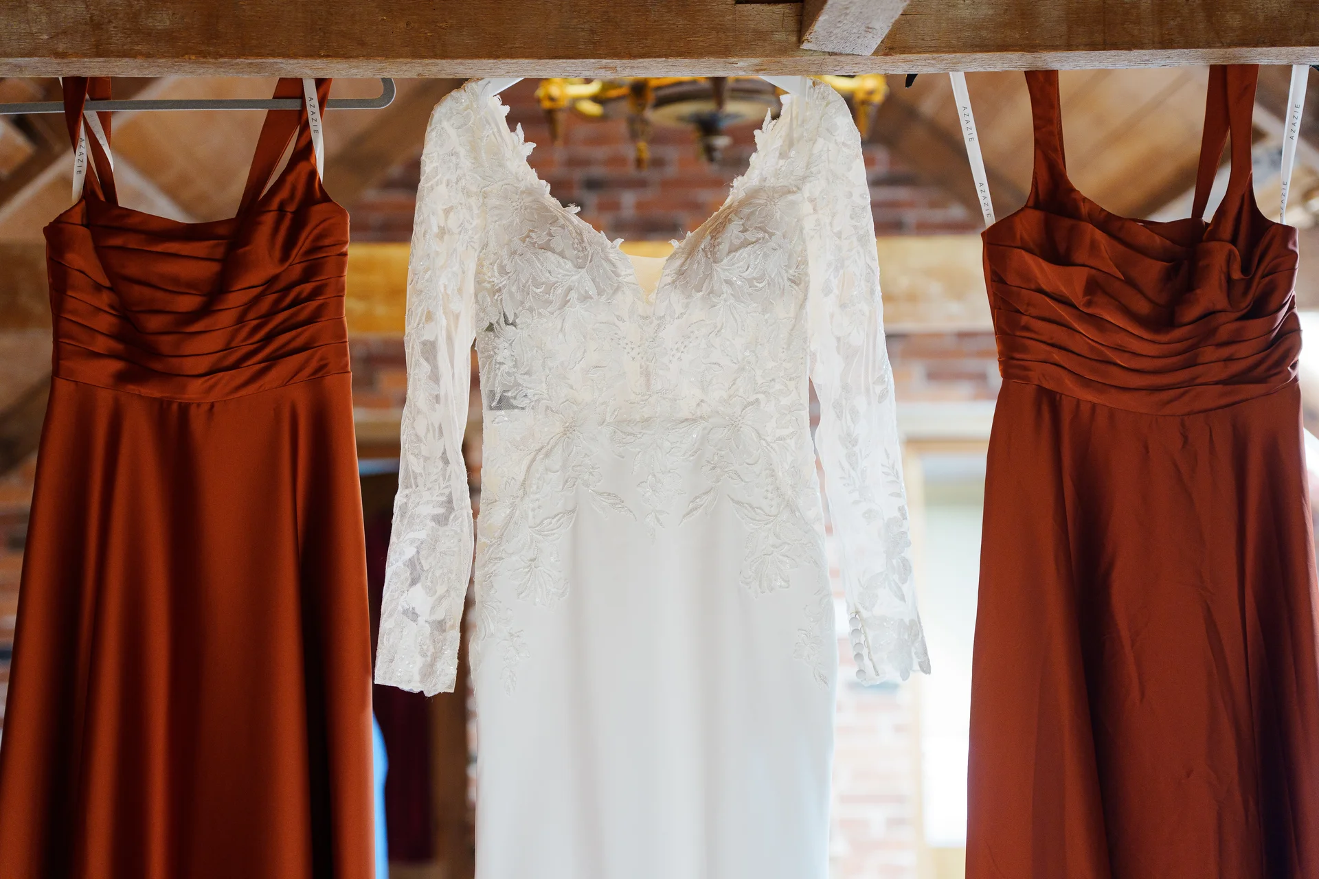 A white lace wedding dress with long sleeves hangs centrally between two rust orange bridesmaid dresses against an exposed brick and wooden beam backdrop.