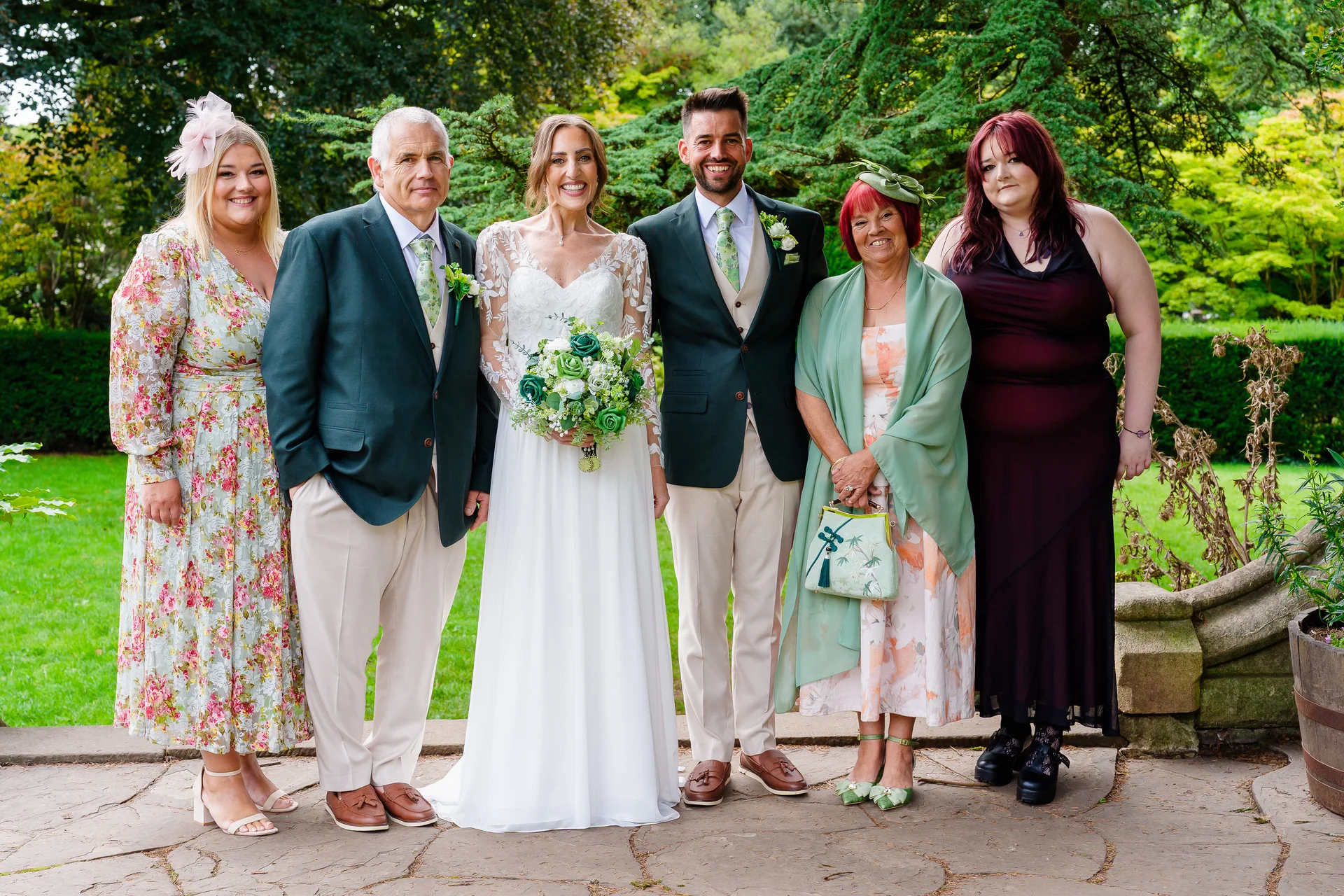 The bride and groom pose outdoors in a manicured garden alongside family members, all dressed in formal attire with subtle green accents.