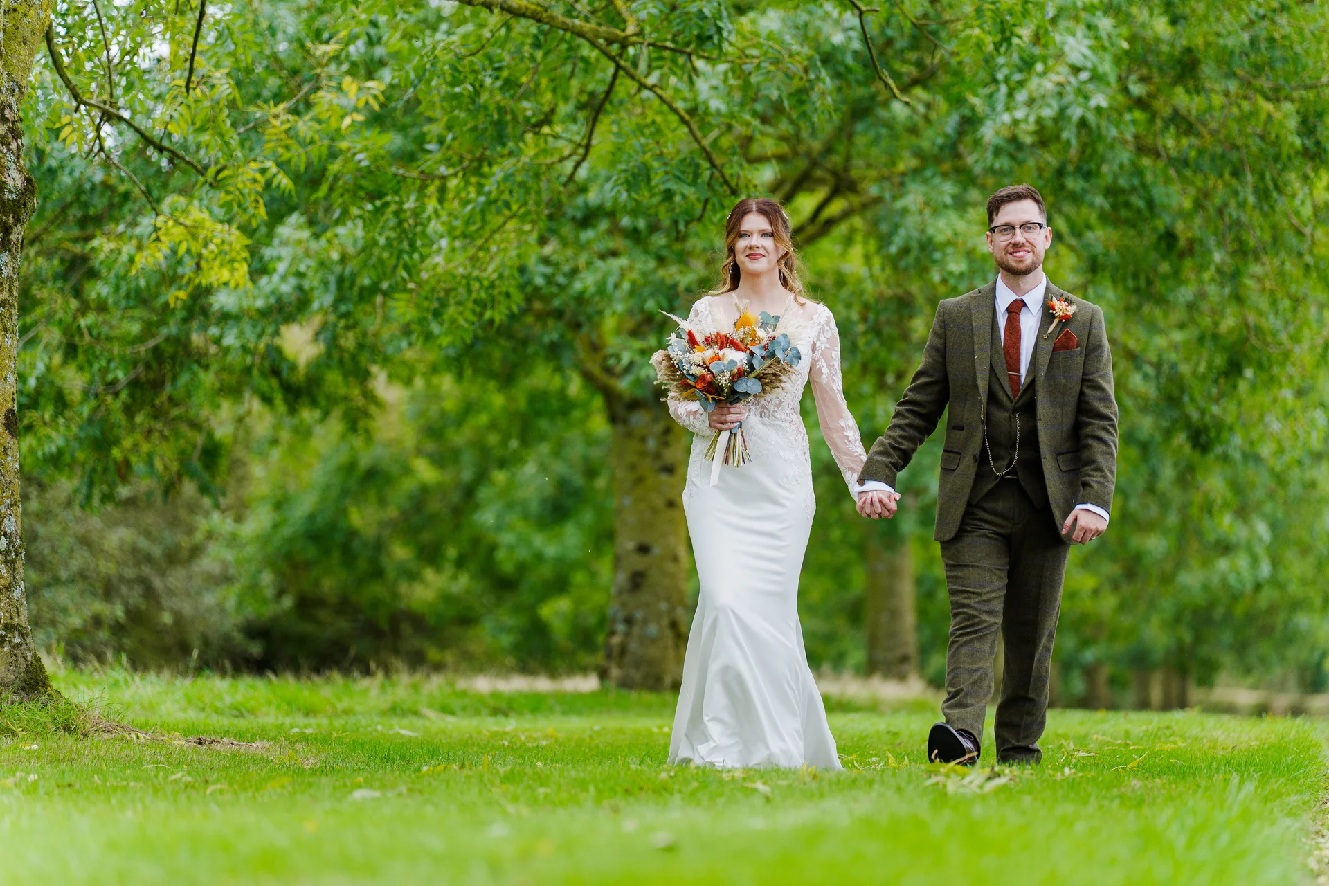 A newlywed couple holds hands and walks through a lush green woodland avenue during their autumn wedding portrait session.