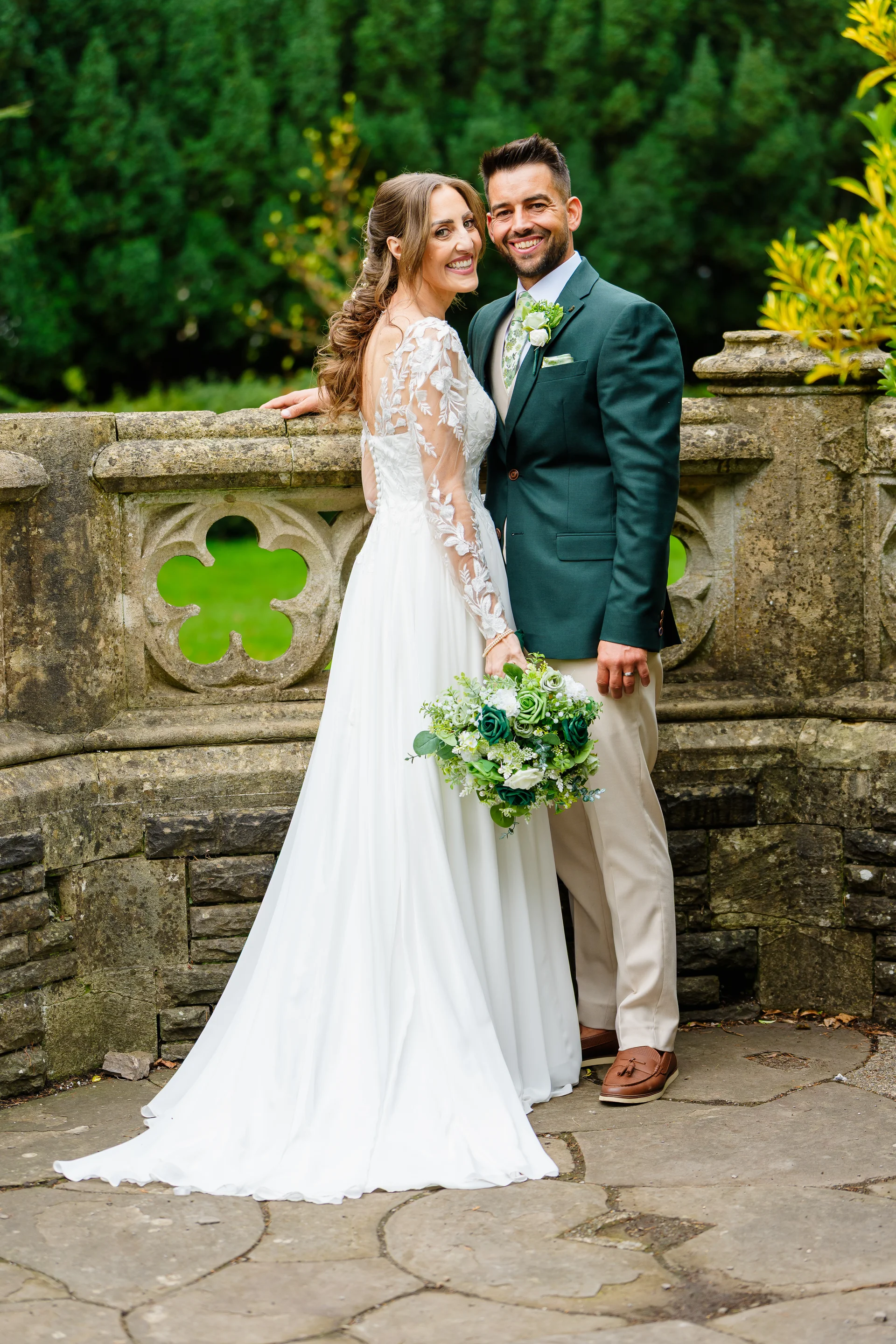 A newlywed couple poses smilingly against an ornate stone balustrade in a lush garden setting, the bride in a long-sleeved lace wedding dress holding a green and white bouquet and the groom in a smart green blazer.