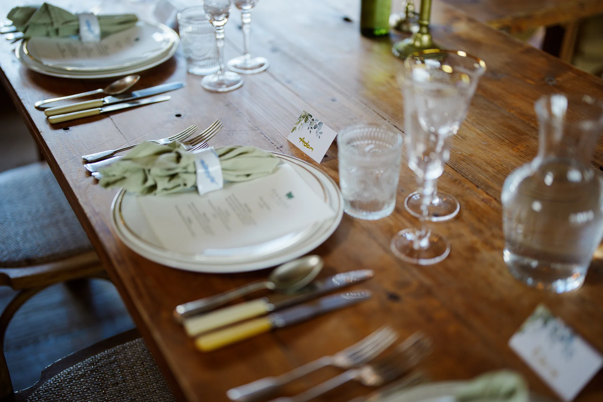 A rustic wooden banquet table is elegantly set with white plates, menu cards, sage green napkins, and delicate crystal glassware.