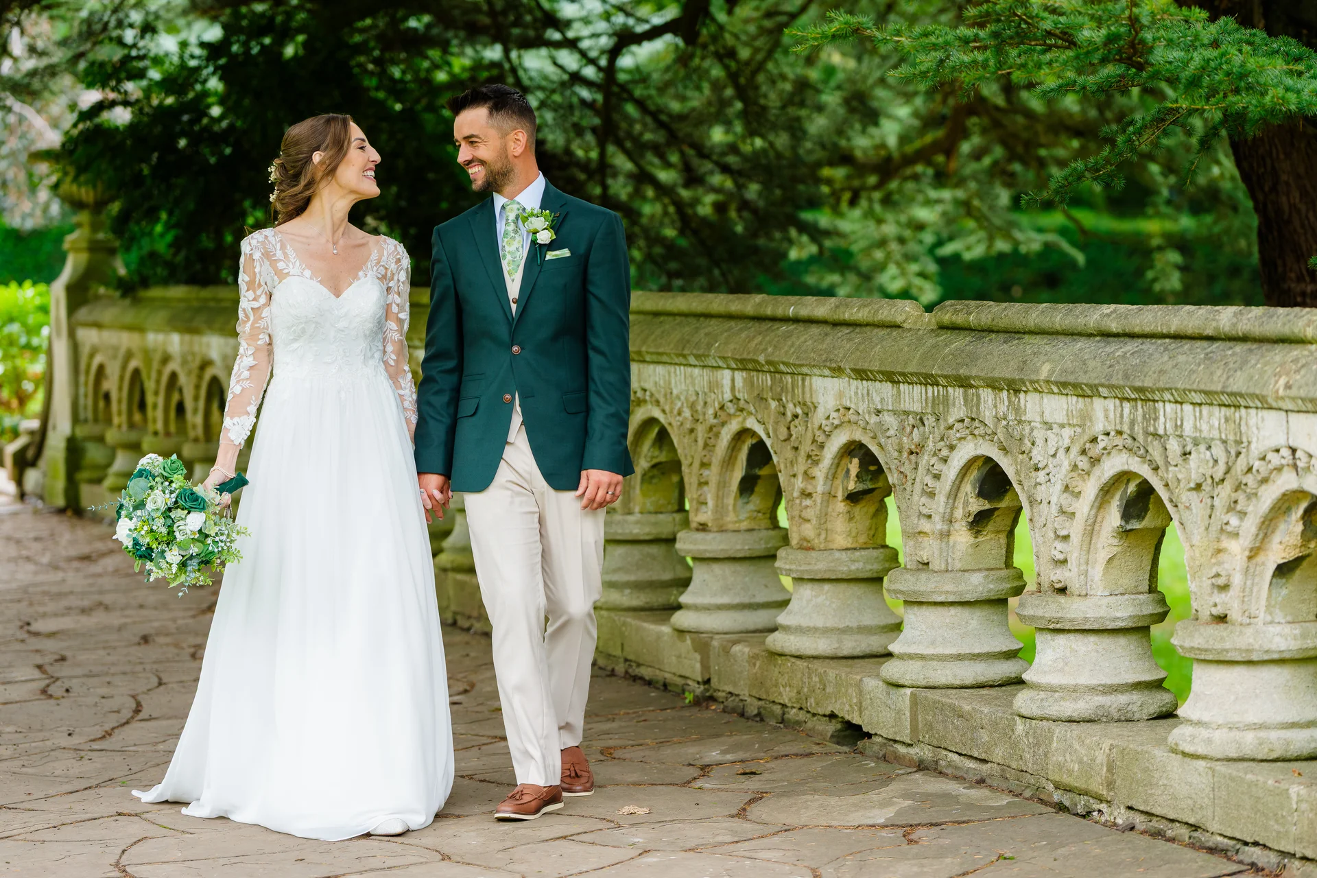 A newly married couple strolls happily together across an ornate stone bridge, the bride holding a lush hand-tied bouquet and wearing an elegant lace-sleeved gown.