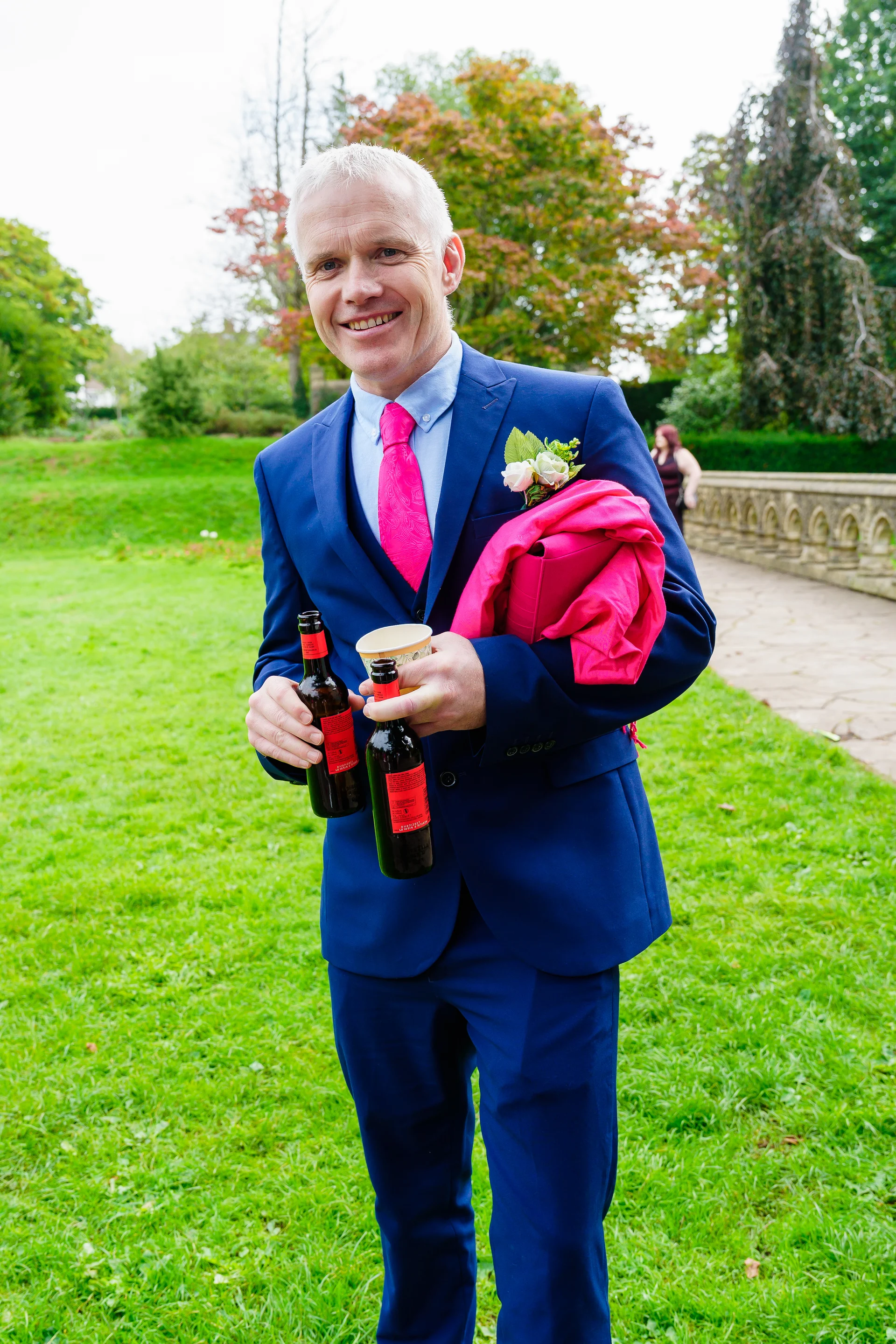 A smiling wedding guest wearing a blue suit and vibrant pink tie stands on a green lawn, holding two bottles of beer and a jacket.